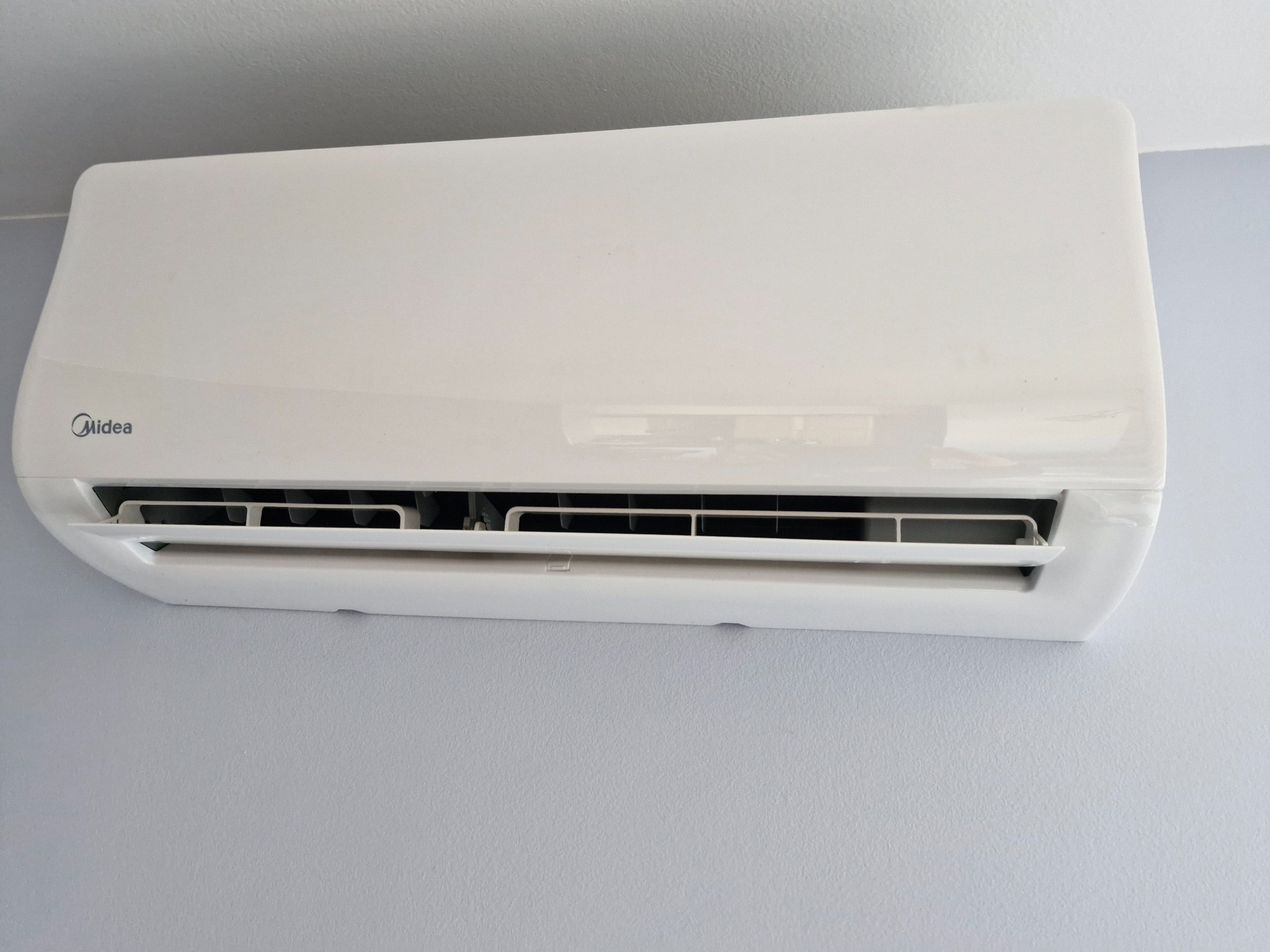 Biały klimatyzator Midea zamontowany na szarej ścianie, widoczne żaluzje w pozycji otwartej.