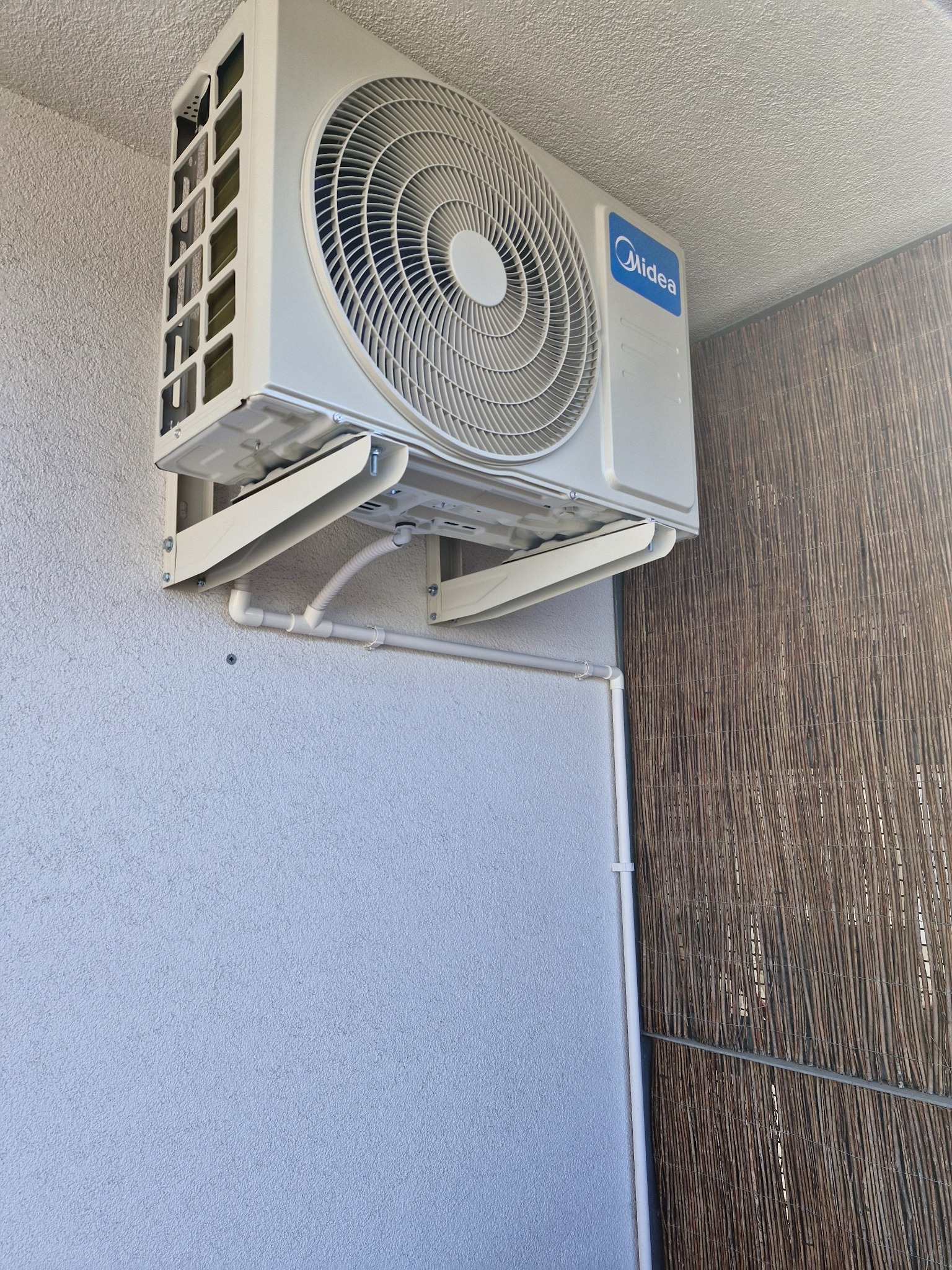 Zewnętrzna jednostka klimatyzacji Midea zamontowana na ścianie z widocznymi rurami odprowadzającymi skropliny, na tle ściany z fakturą i fragmentu bambusowej maty.