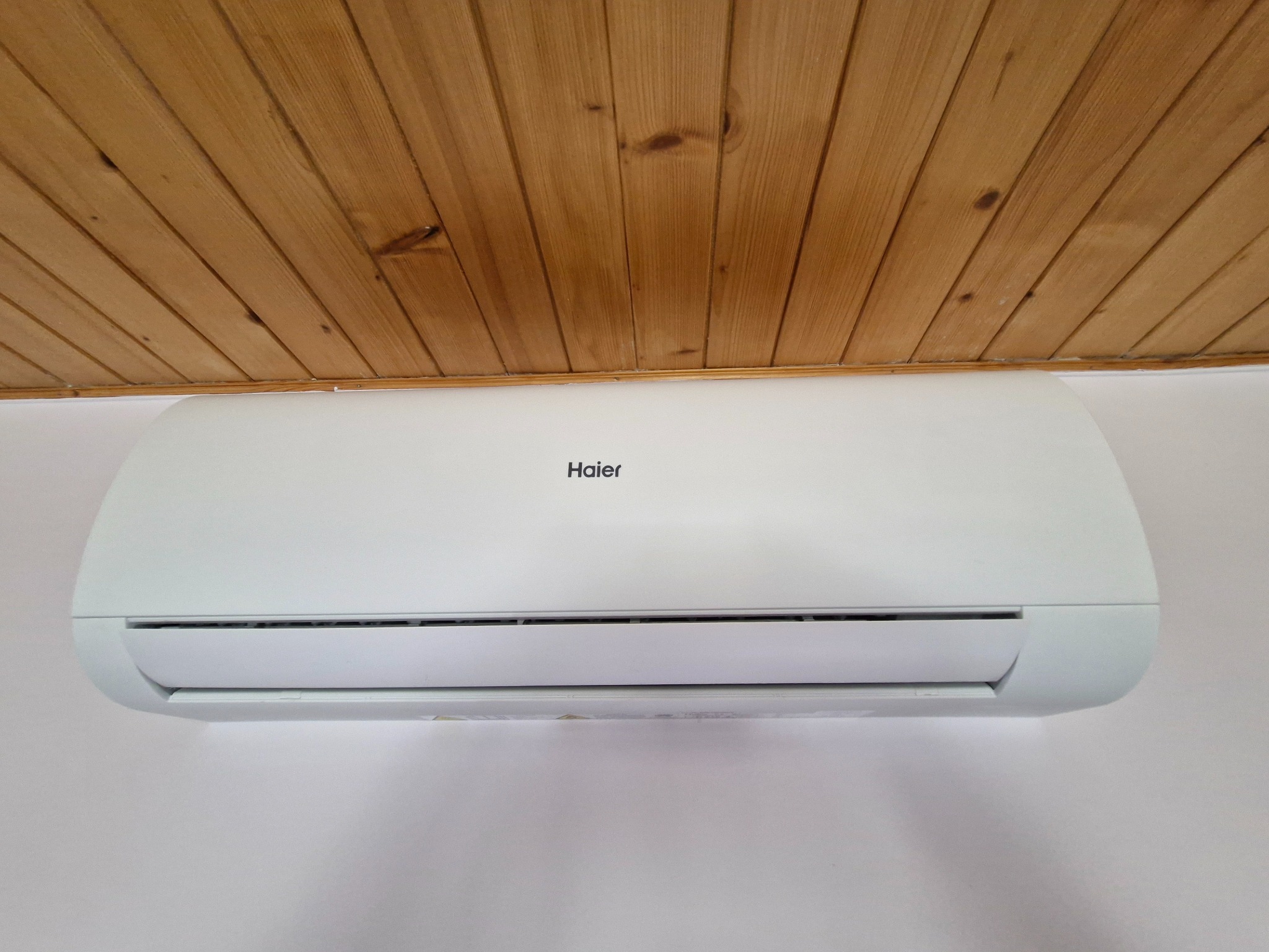 Biały klimatyzator Haier zamontowany na białej ścianie pod drewnianym sufitem z boazerii.