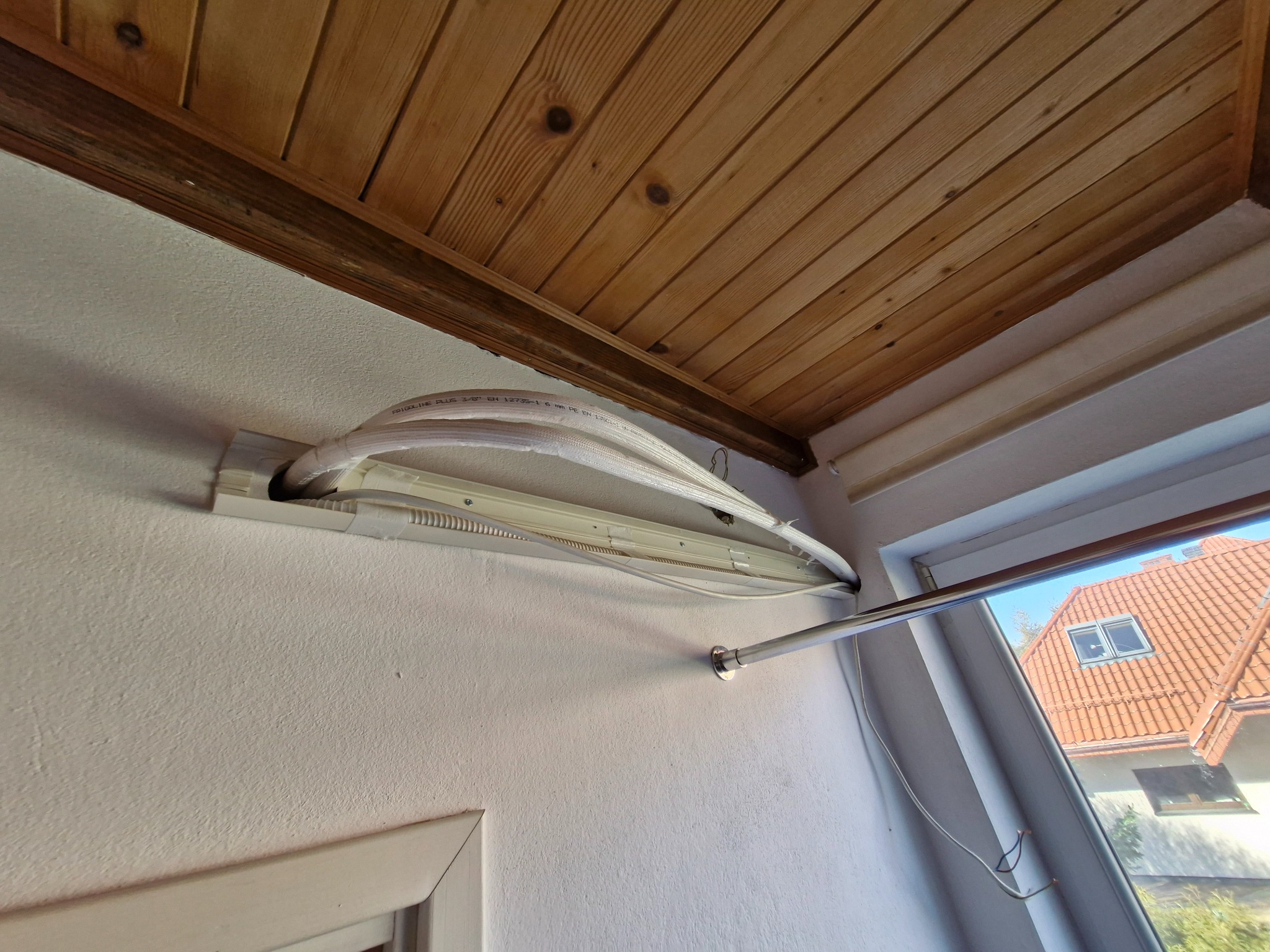 Białe rury instalacyjne wystające ze ściany pod drewnianym sufitem, widok z dołu, fragment okna z widokiem na dom z czerwoną dachówką.