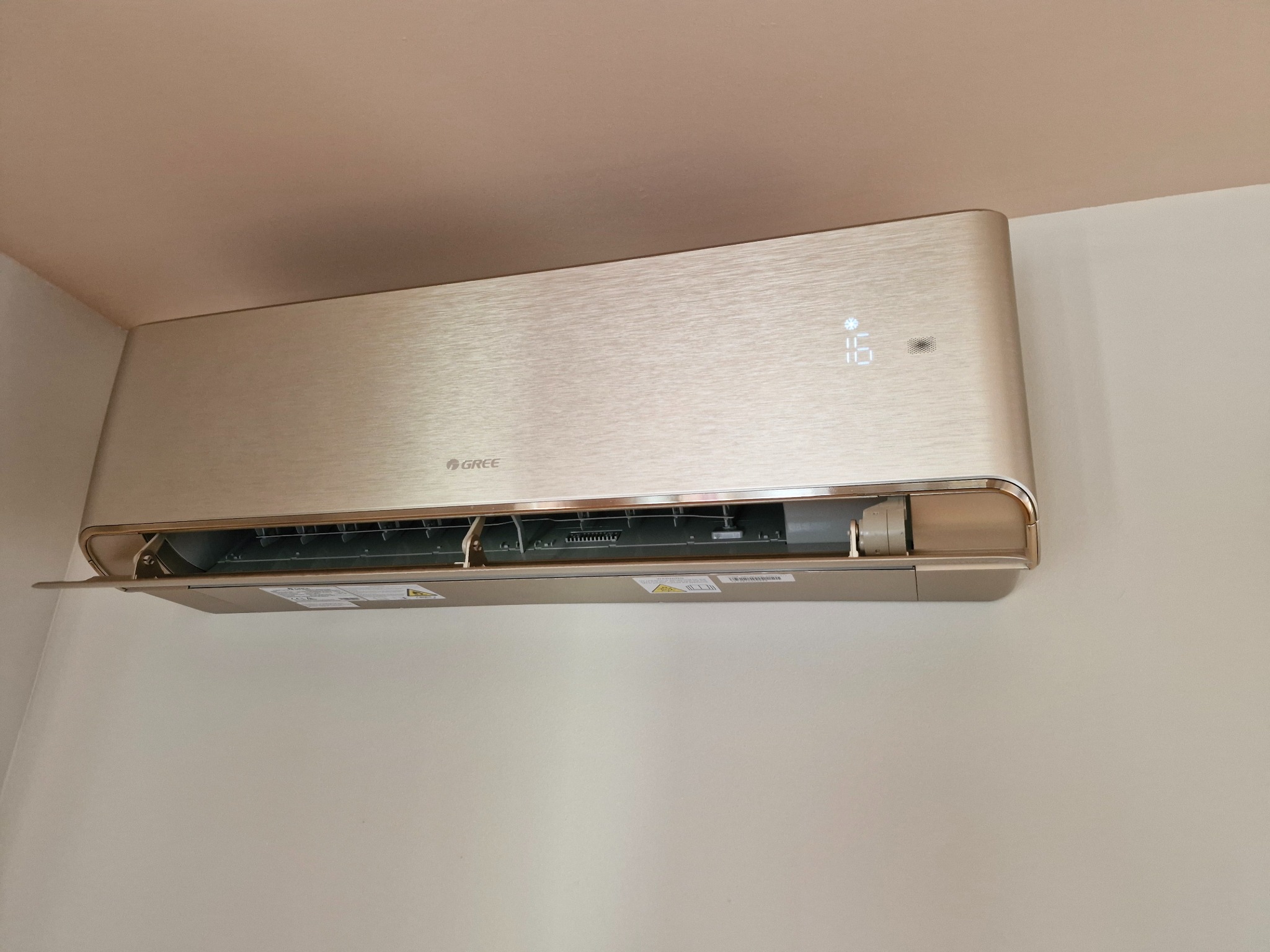 Złoty klimatyzator marki Gree zamontowany na białej ścianie, z wyświetlaczem wskazującym temperaturę 16 stopni Celsjusza.