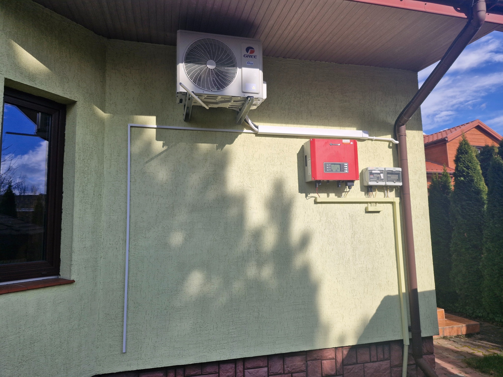 Zewnętrzna jednostka klimatyzatora Gree zamontowana na elewacji domu, obok skrzynki elektrycznej i falownika fotowoltaicznego.
