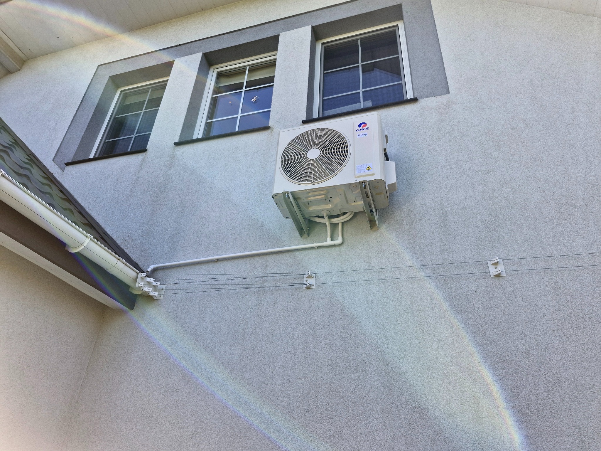 Biały klimatyzator Gree zamontowany na elewacji domu, widoczny z boku, z trzema oknami w tle i fragmentem rynny. Na ścianie tęczowa poświata.