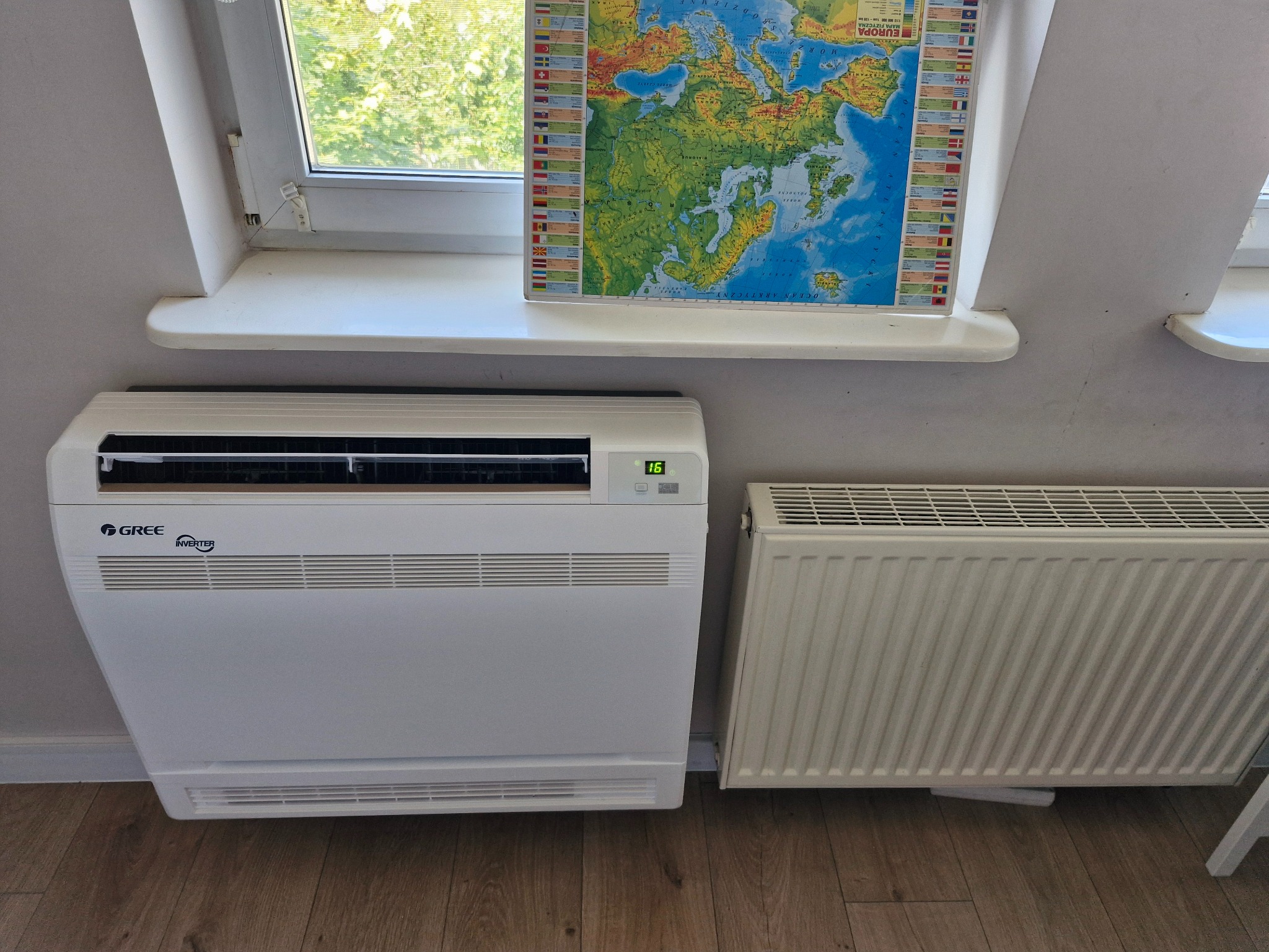 Biały klimatyzator pod oknem na poddaszu, obok grzejnik. Na parapecie mapa Europy. Widoczne logo Gree Inverter. Ustawiona temperatura 16 stopni.