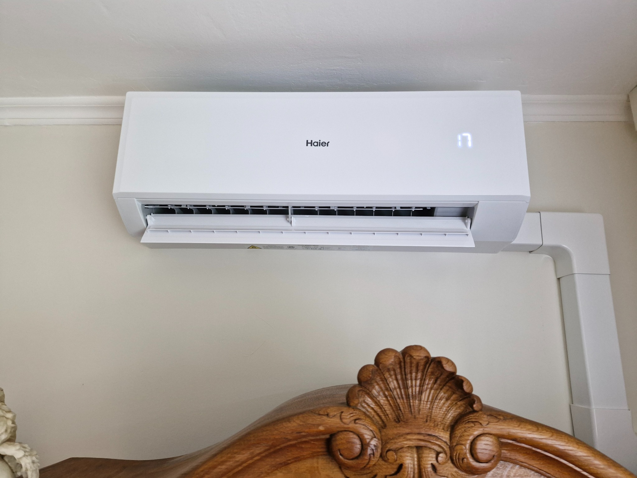 Biała klimatyzacja Haier na ścianie nad drewnianym zagłówkiem łóżka. Widoczny wyświetlacz z temperaturą 17 stopni Celsjusza.