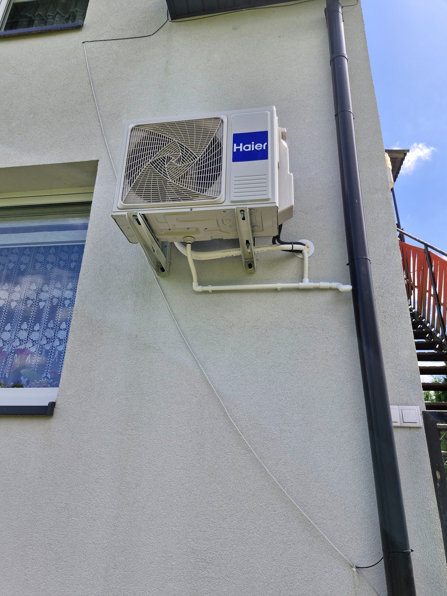 Biały klimatyzator Haier zamontowany na elewacji budynku obok okna z firanką i rury spustowej. Widoczne białe rurki instalacji klimatyzacji.