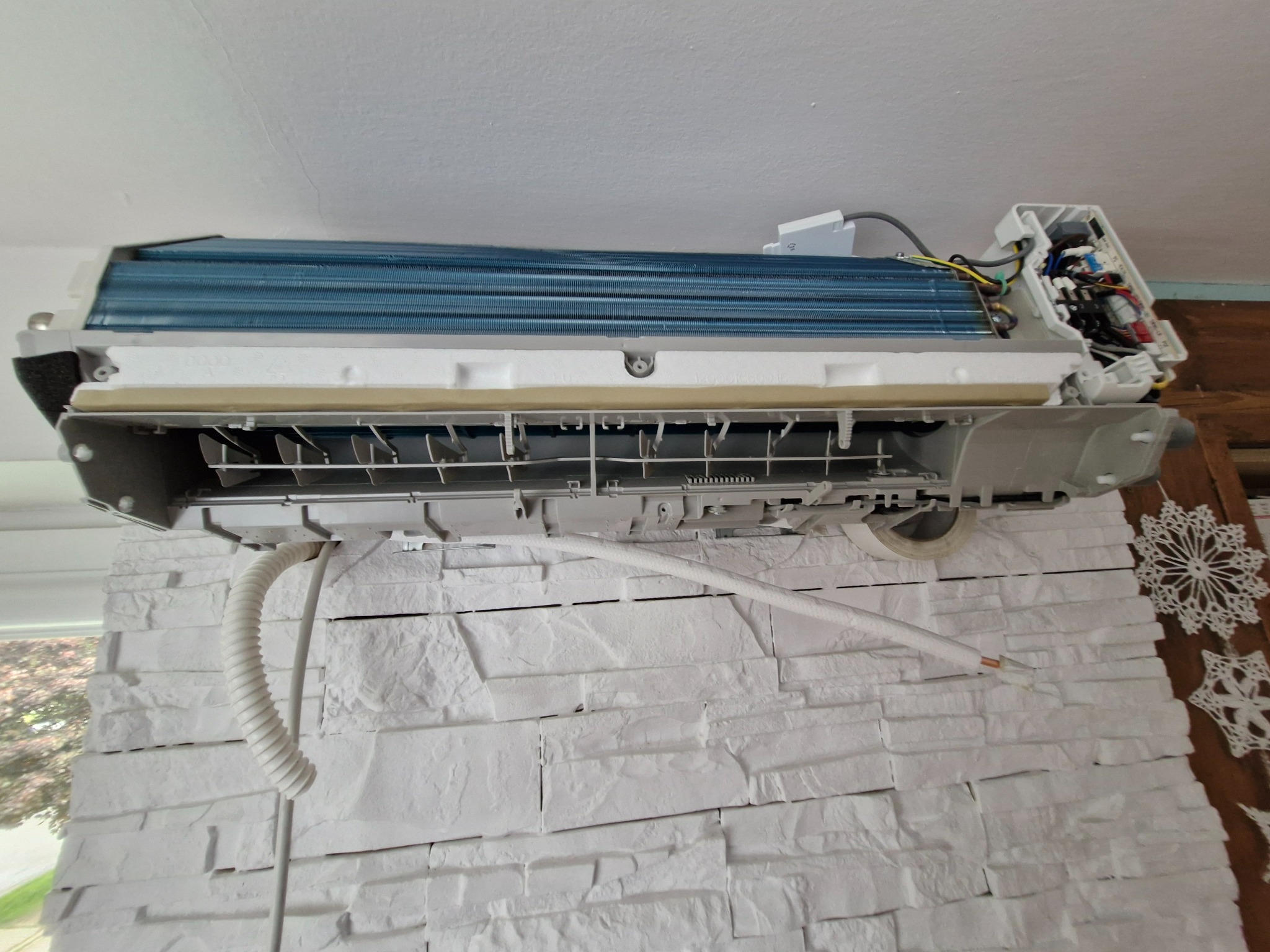 Rozłożony klimatyzator ścienny z widocznymi elementami wewnętrznymi, zamontowany na dekoracyjnej ścianie z białej cegły. Widoczne przewody i elementy instalacji.