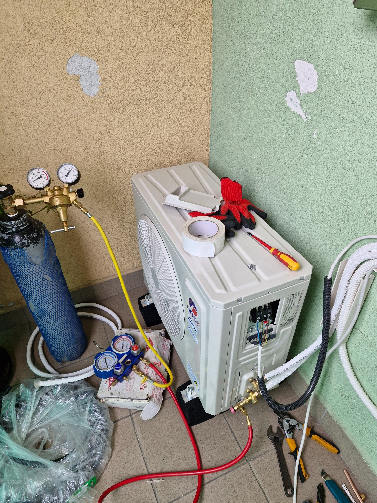 Montaż klimatyzatora: butla z gazem, manometry, narzędzia, przewody i rękawice obok jednostki zewnętrznej na balkonie z zielono-beżową ścianą.