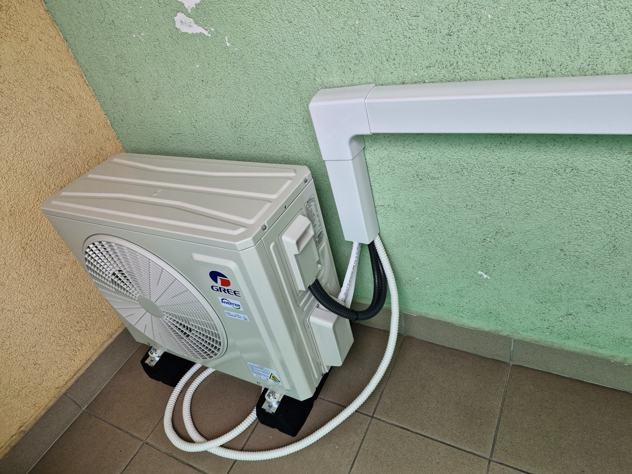 Zamontowany klimatyzator Gree na zewnątrz budynku, z widocznymi przewodami i elementami instalacji. Ściana w kolorze zielonym i beżowym, brązowe płytki podłogowe.