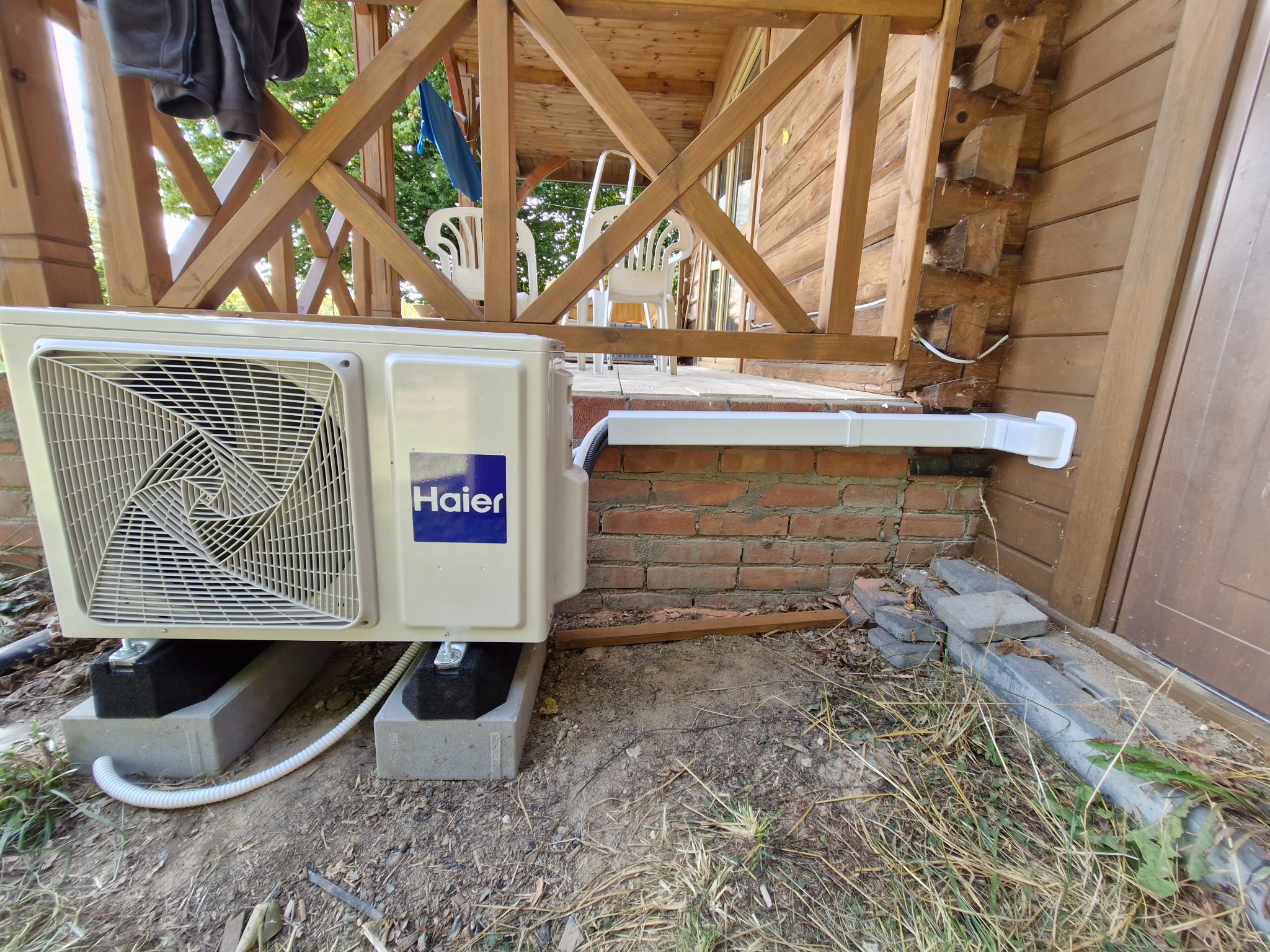 Zewnętrzna jednostka klimatyzacji Haier zamontowana na betonowych blokach przy drewnianym budynku. Widoczna instalacja odprowadzająca skropliny.