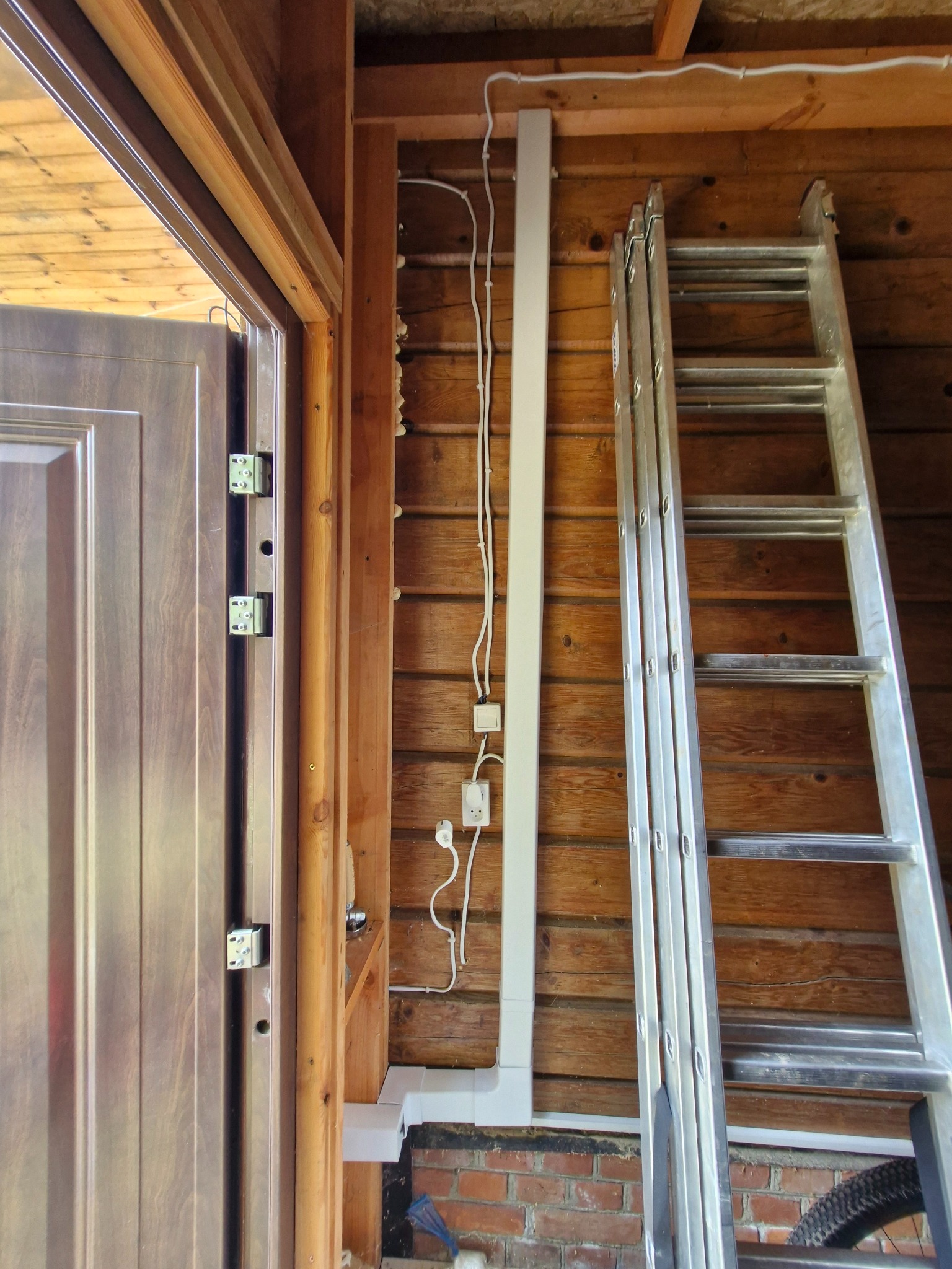 Montaż instalacji elektrycznej w drewnianym domu: białe korytko z przewodami i gniazdkami, drabina oparta o ścianę, widok od strony drzwi.
