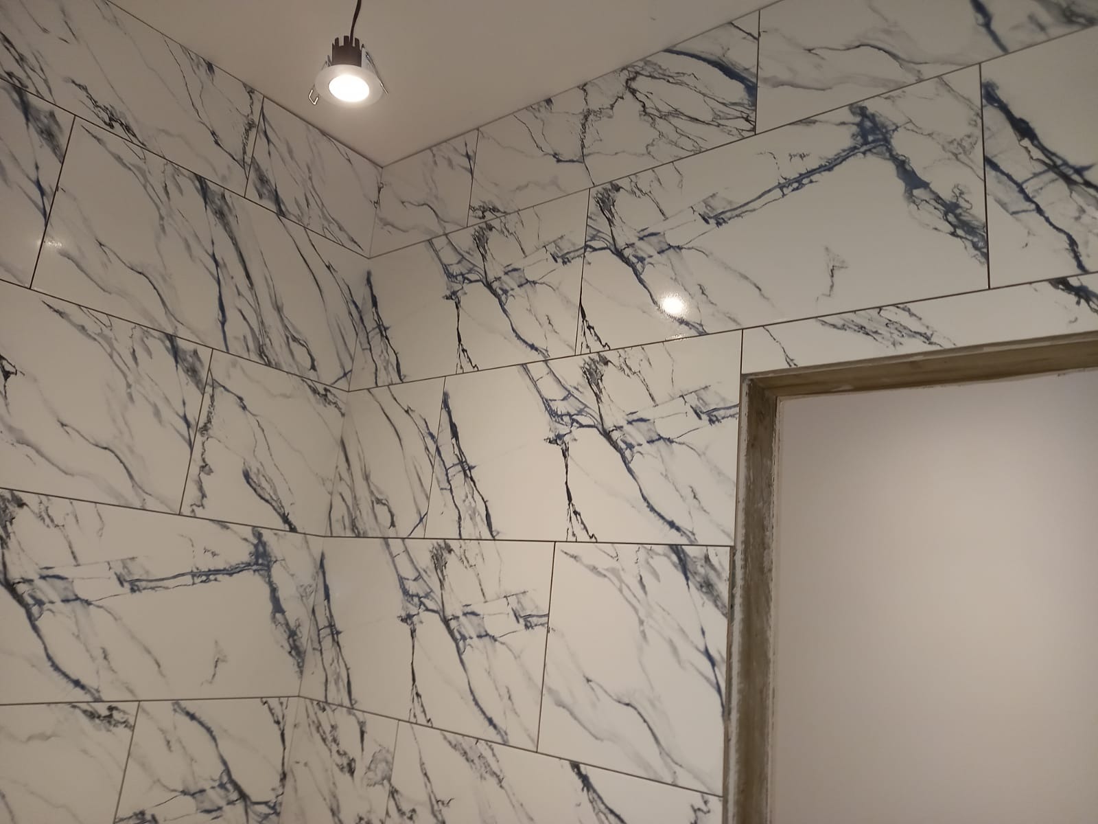 Ściana i narożnik wyłożone białymi płytkami imitującymi marmur z wyraźnym, ciemnym żyłkowaniem; widoczny fragment oświetlenia sufitowego i surowa ościeżnica.
