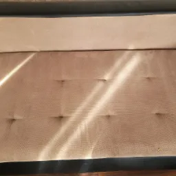 Fragment beżowej kanapy z ciemnymi elementami, widoczne wgniecenia i plamka na siedzisku, oświetlone promieniami słońca