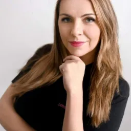 Portret uśmiechniętej logopedki z dłonią pod brodą, w czarnej bluzce z dyskretnym, różowym napisem 'LOGO'.