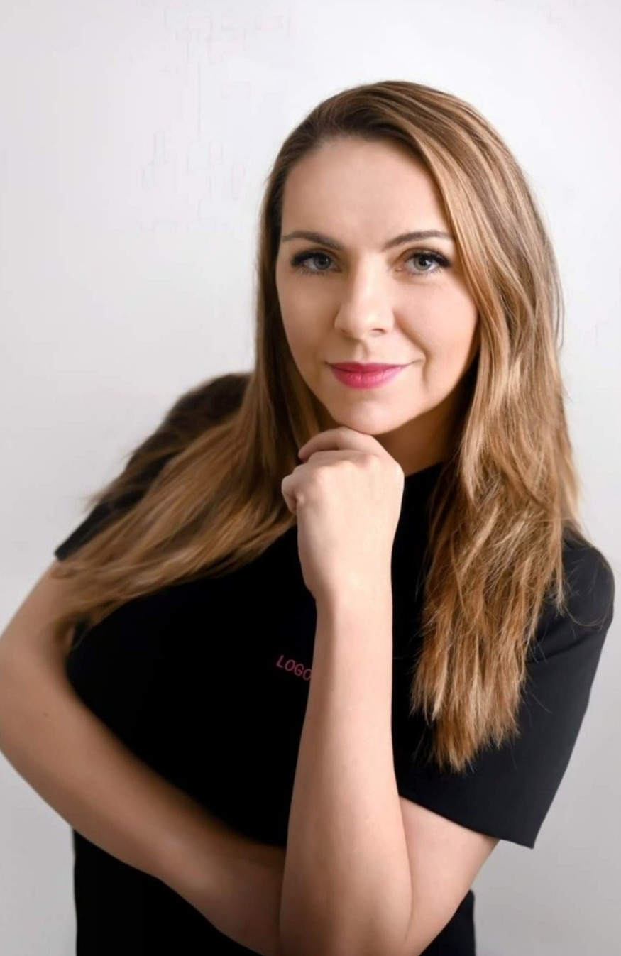 Portret uśmiechniętej logopedki z dłonią pod brodą, w czarnej bluzce z dyskretnym, różowym napisem 'LOGO'.