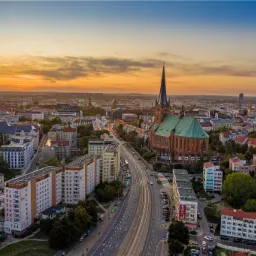 Panoramiczny widok z lotu ptaka na miasto o zachodzie słońca, z dominującą gotycką katedrą z zielonym dachem i wysoką wieżą, szeroką arterią komunikacyjną oraz zabudową mieszkalną w tle...