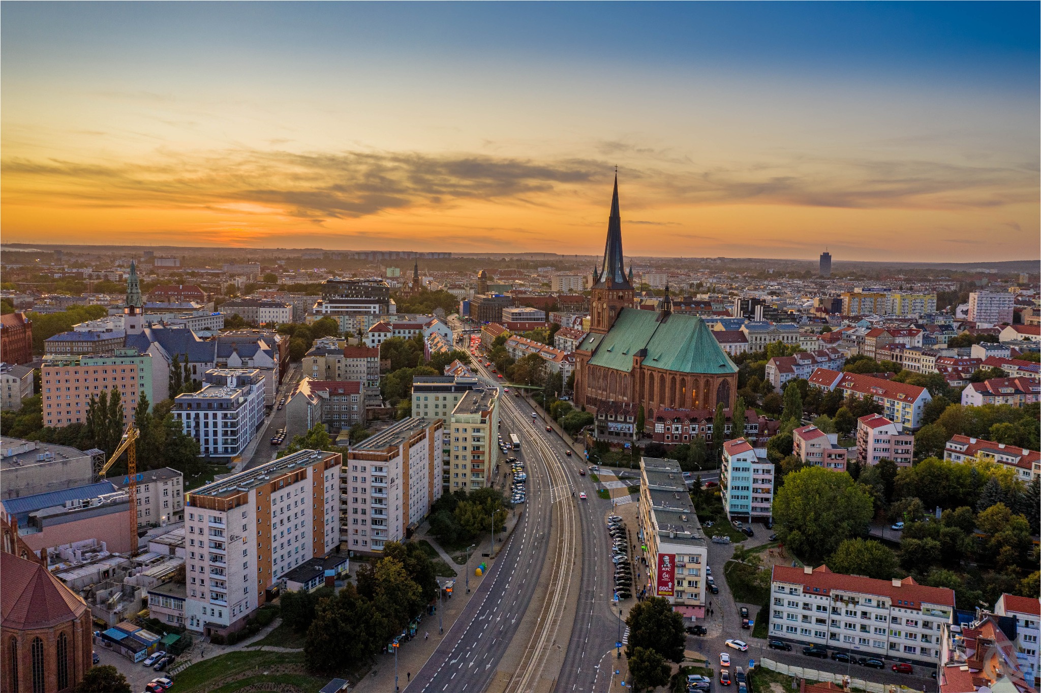 Panoramiczny widok z lotu ptaka na miasto o zachodzie słońca, z dominującą gotycką katedrą z zielonym dachem i wysoką wieżą, szeroką arterią komunikacyjną oraz zabudową mieszkalną w tle...