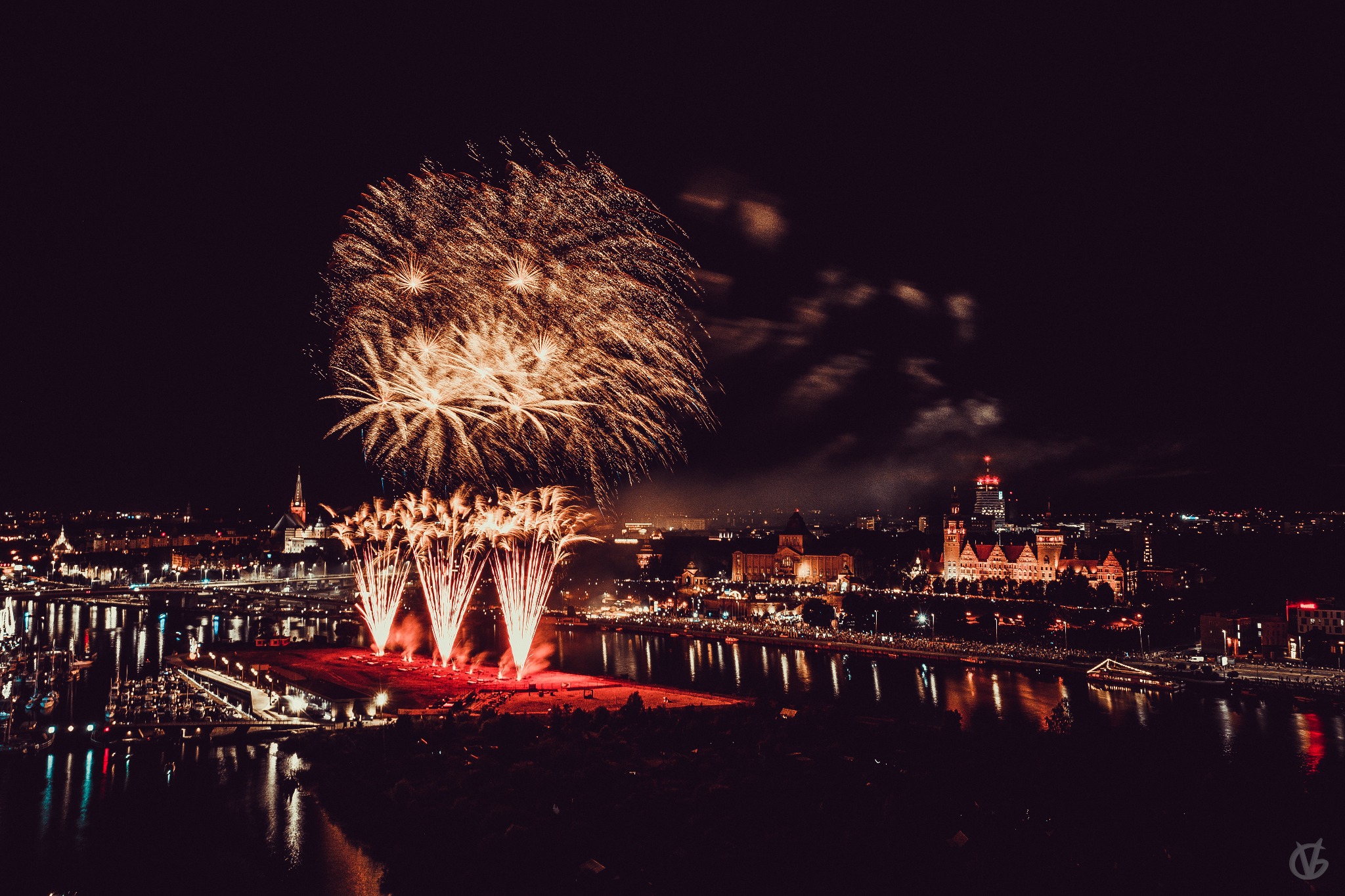 Nocny widok na panoramę miasta z rozbłyskami fajerwerków nad rzeką; odbicia świateł w wodzie.