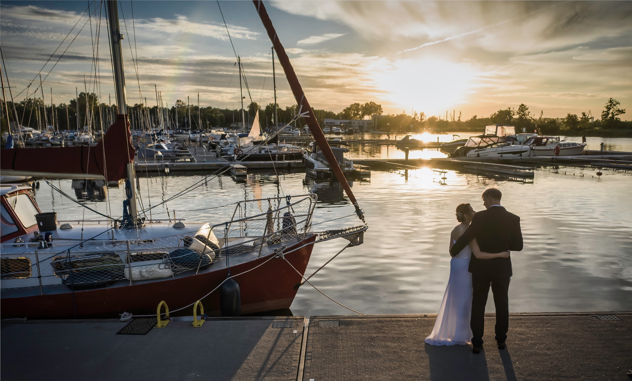 Para młoda obejmująca się na pomoście przy zacumowanej czerwonej żaglówce, w tle marina i zachodzące słońce.