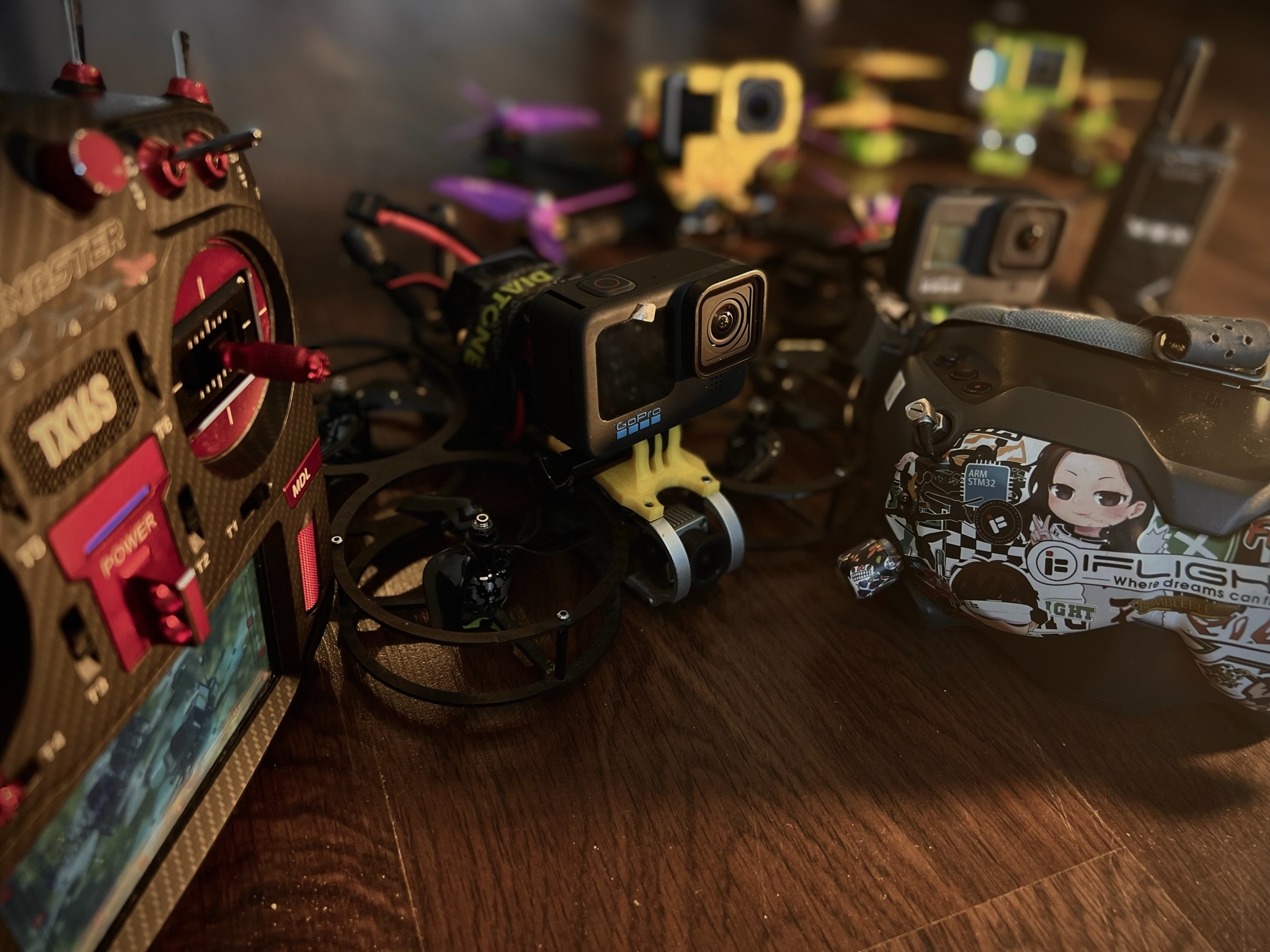 Kolekcja dronów FPV z kamerami GoPro, kontrolerem TX16S i goglami FPV z naklejkami na drewnianej powierzchni.