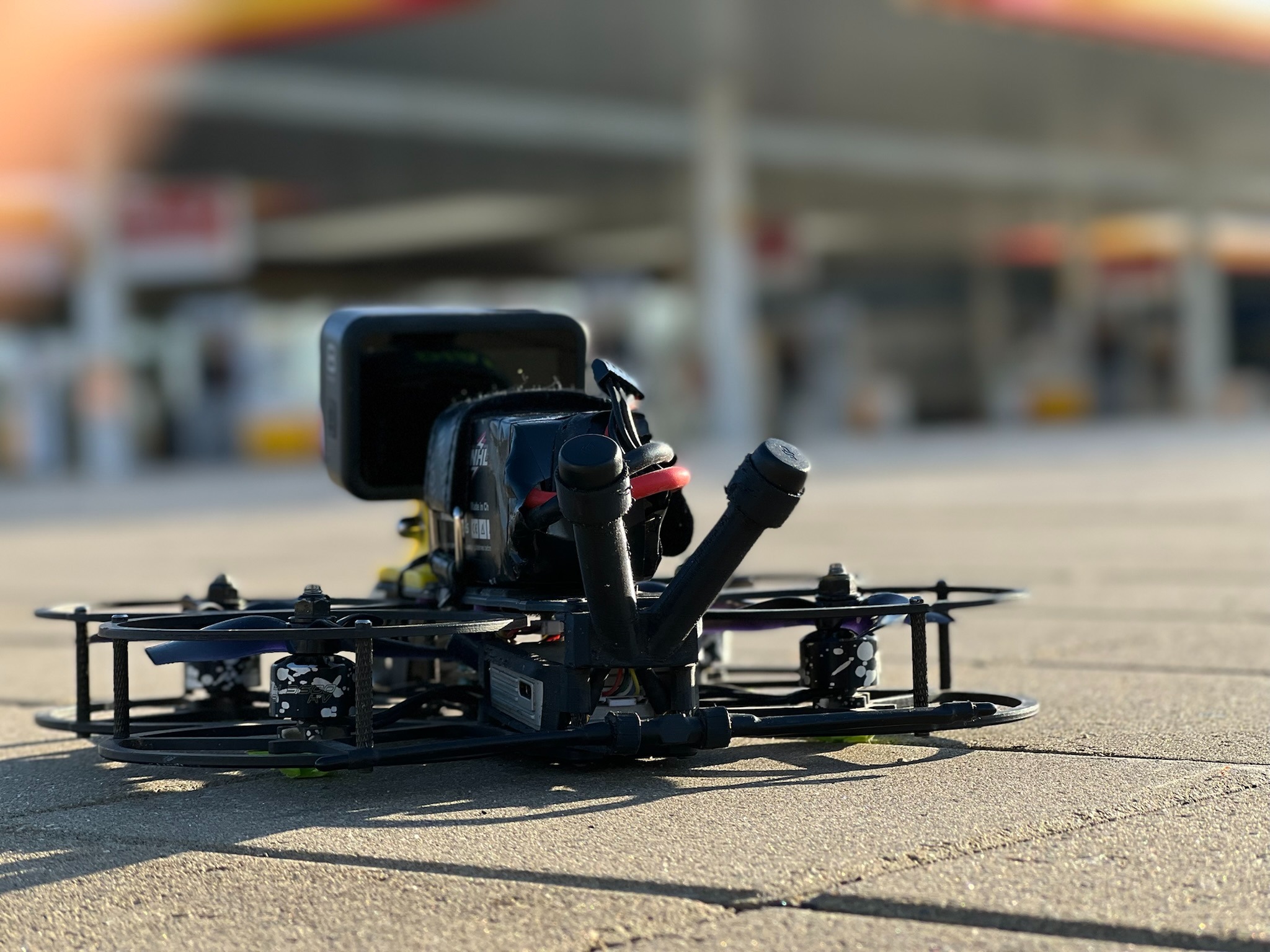 Niski kąt ujęcia drona FPV z kamerą GoPro, stojącego na betonowej powierzchni z rozmytą stacją benzynową w tle.