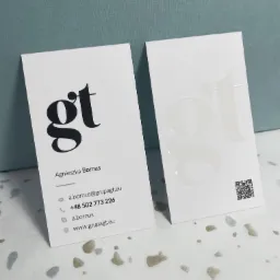 Dwie wizytówki z logo 'gt', jedna z czarnym nadrukiem, druga z tłoczonym, błyszczącym logo, leżące na wzorzystym tle.