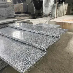 Polerowane płyty z szarego granitu, ułożone warstwowo w warsztacie kamieniarskim, z widoczną piłą do cięcia kamienia w tle.