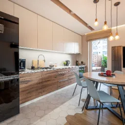 Nowoczesna, jasna kuchnia z drewnianymi szafkami, okrągłym stołem i krzesłami w odcieniach szarości, z widokiem na balkon z roślinami, oświetlona wiszącymi lampami z żarówkami Edisona.