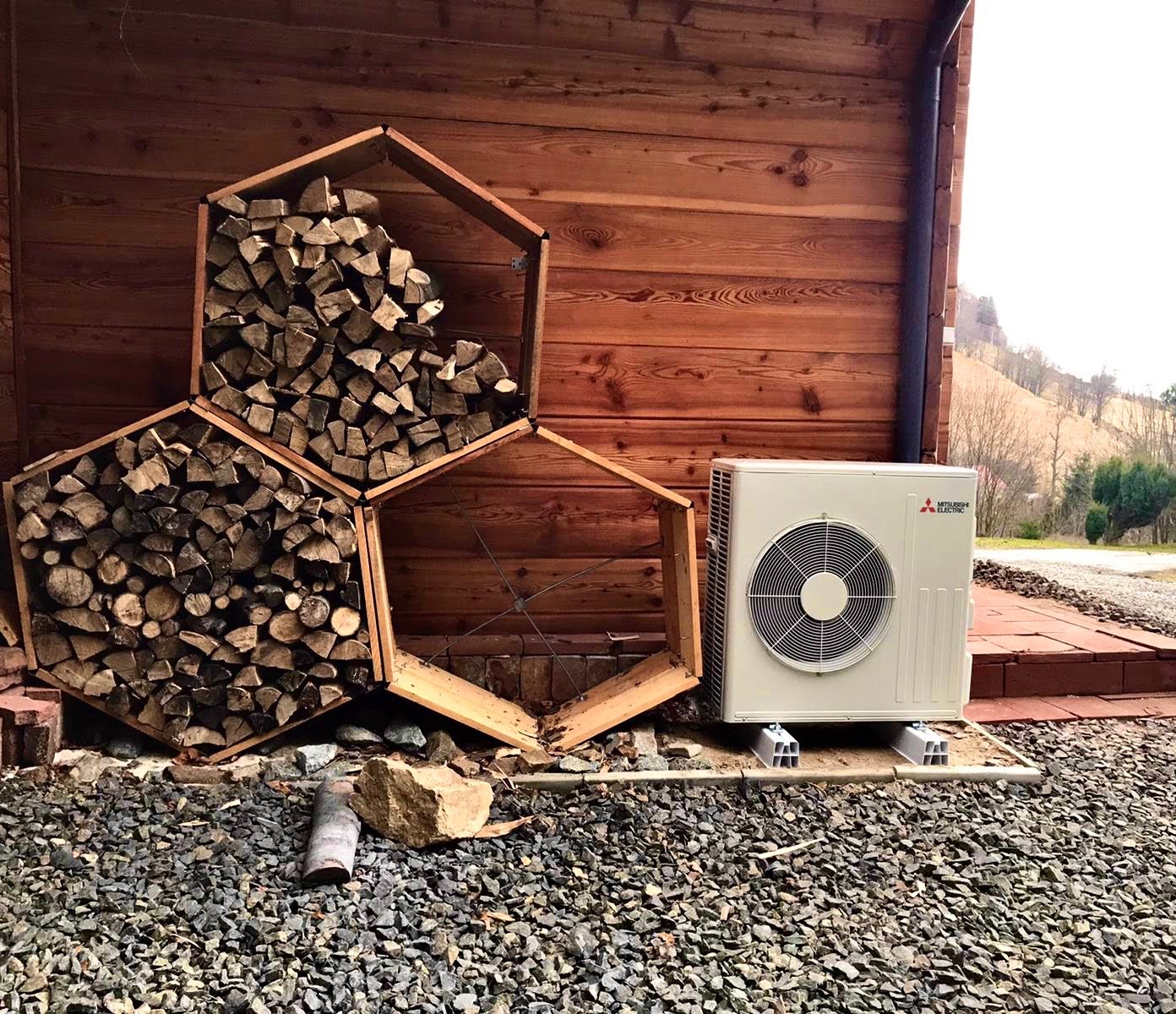 Zewnętrzna jednostka klimatyzacji Mitsubishi Electric na betonowych podstawach, obok drewnianej ściany i heksagonalnych półek na drewno, częściowo wypełnionych. Widoczny kamienisty teren wokół.