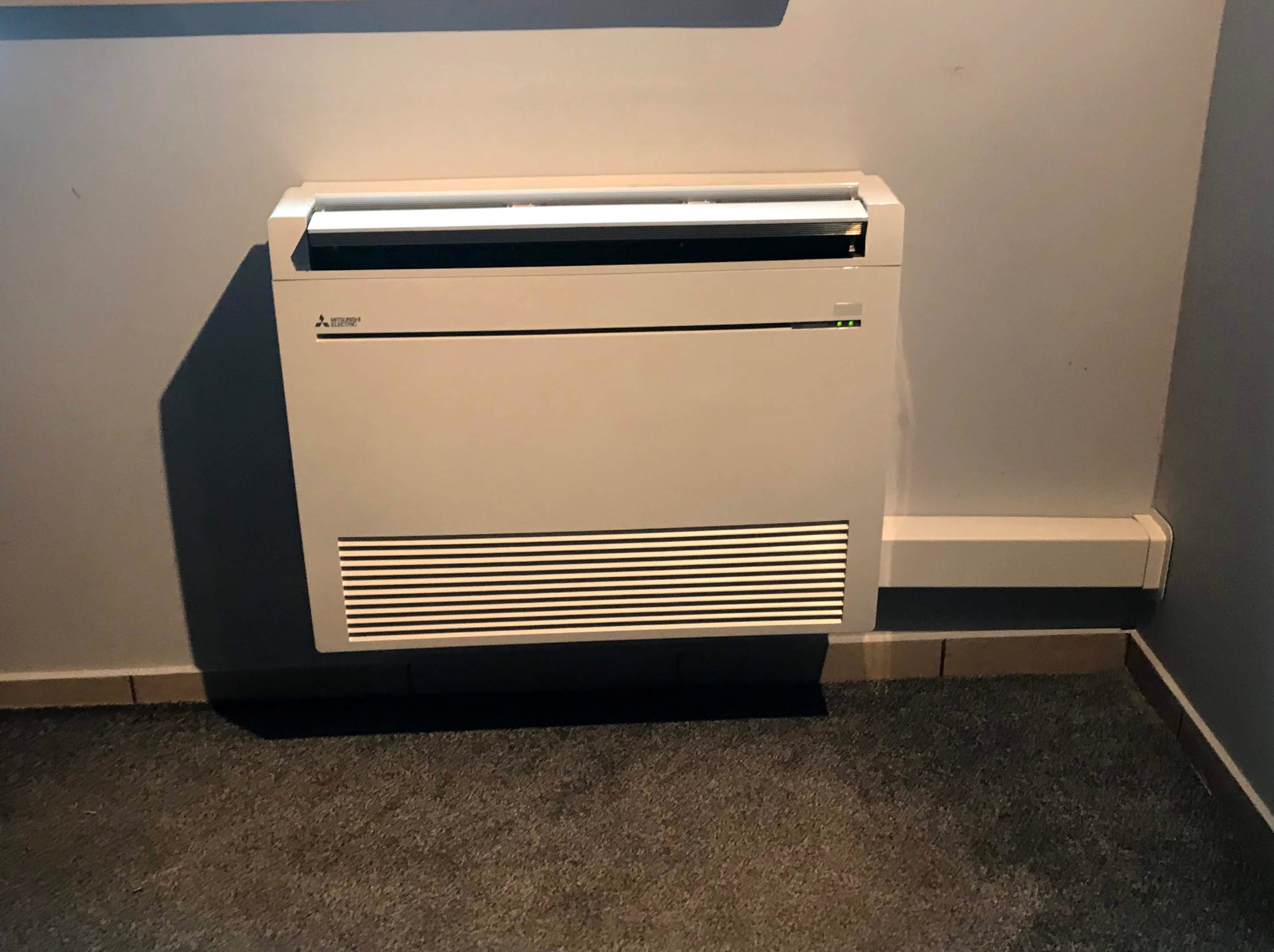 Klimatyzator przypodłogowy Mitsubishi Electric w jasnej obudowie, zamontowany na szarej ścianie w pomieszczeniu z ciemną wykładziną.