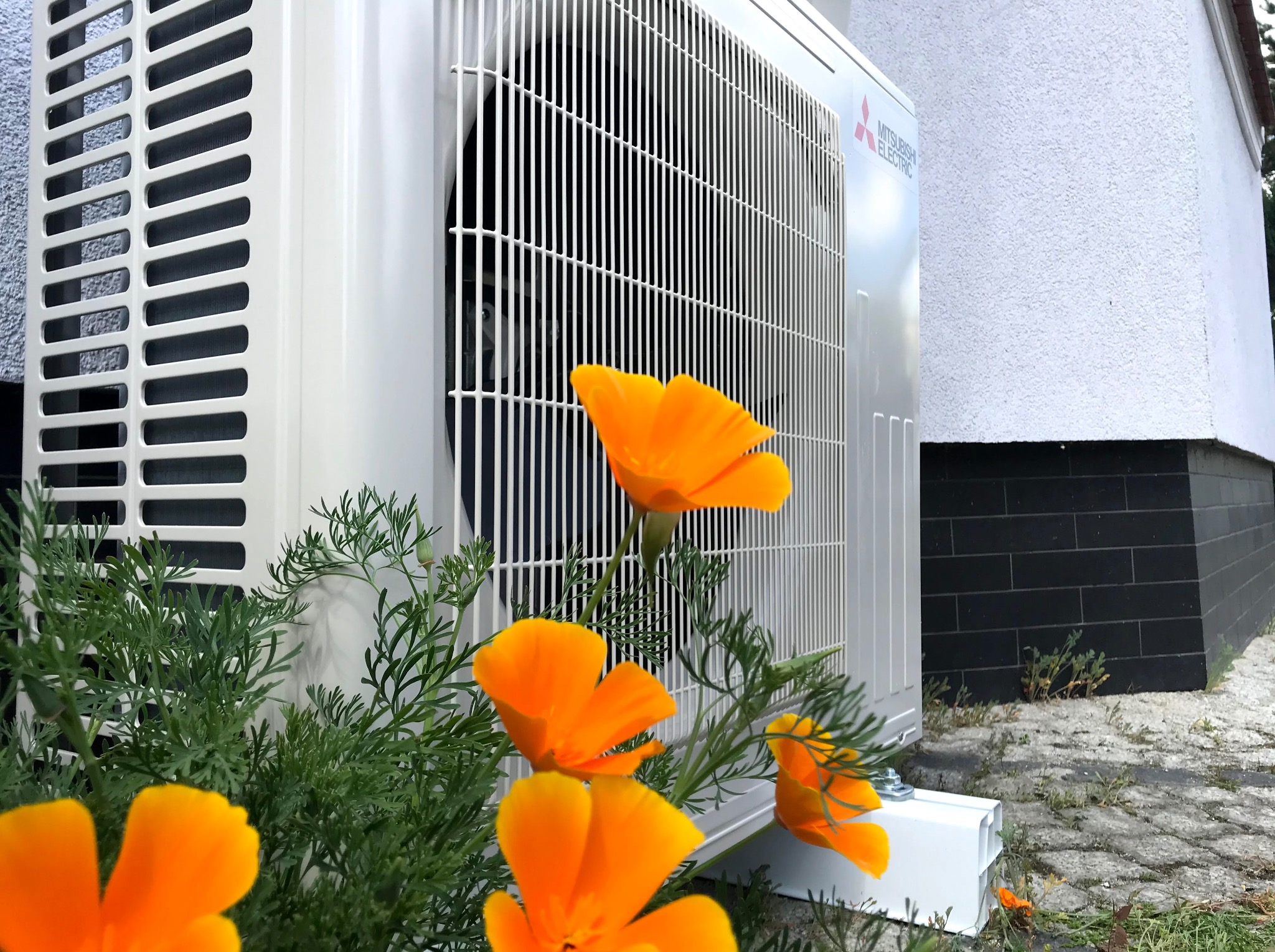 Zewnętrzna jednostka klimatyzacji Mitsubishi Electric, zamontowana na elewacji budynku, otoczona kwitnącymi, pomarańczowymi kwiatami kalifornijskimi.