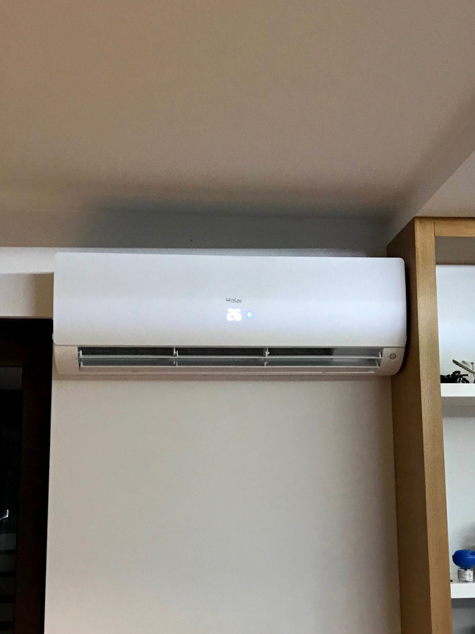 Biała jednostka klimatyzacyjna Haier zamontowana na ścianie w pomieszczeniu, z wyświetlaczem LED wskazującym temperaturę 26 stopni Celsjusza. Obok widoczny fragment regału.