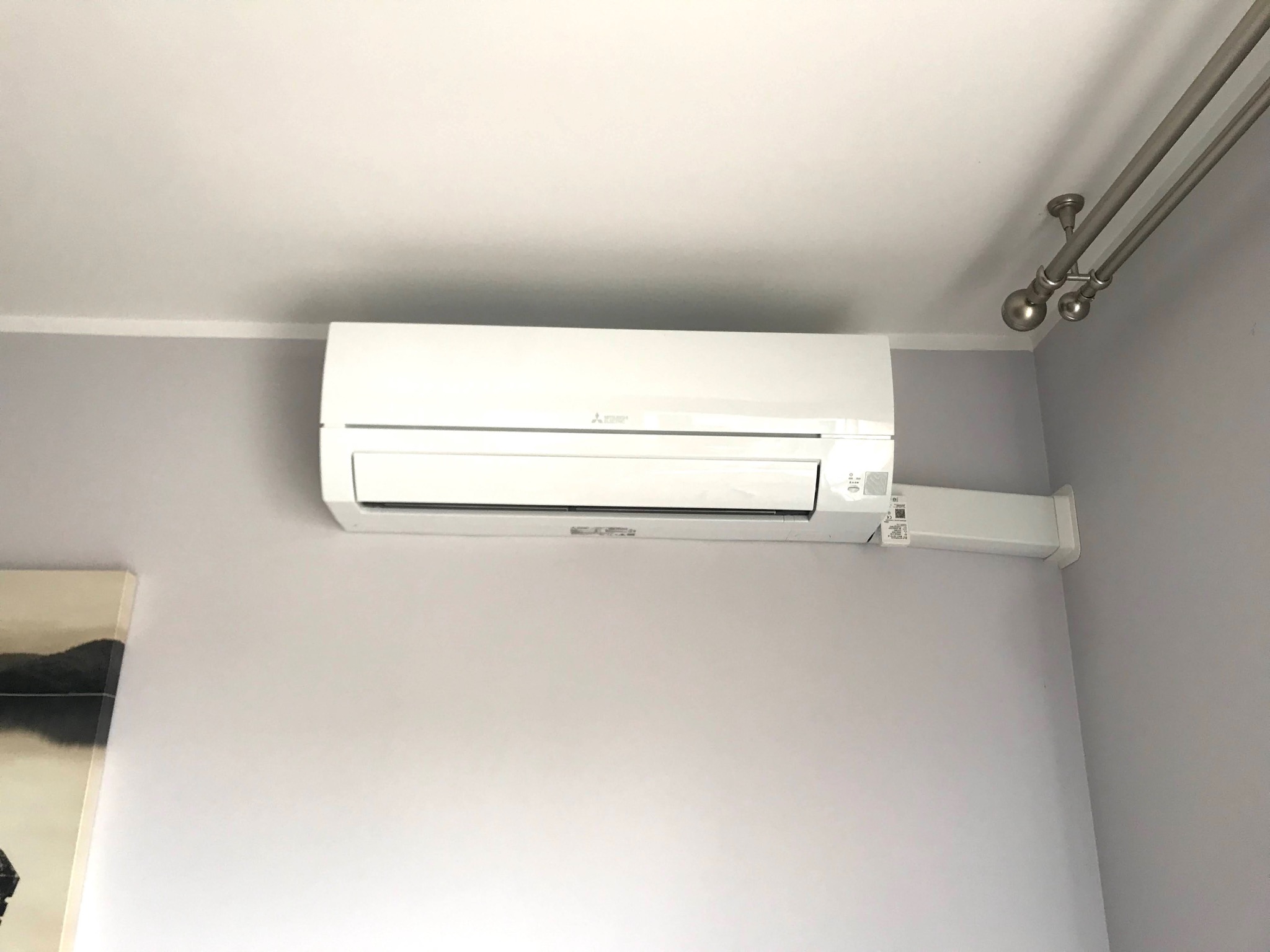 Biała jednostka klimatyzacji Mitsubishi zamontowana na szarej ścianie w pomieszczeniu, widoczny fragment obrazu po lewej stronie i karnisz z kulami na suficie.