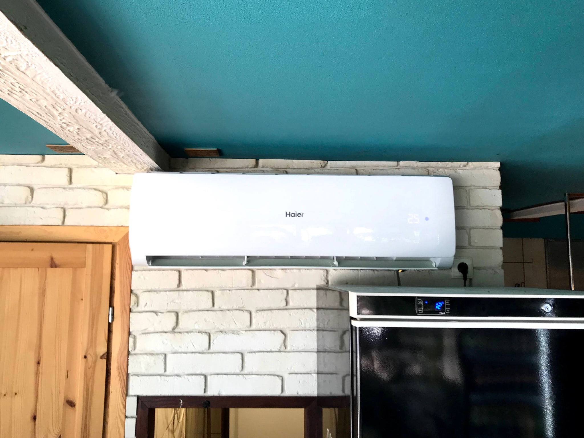 Biały klimatyzator Haier zamontowany na ścianie z białej cegły, widoczny fragment drewnianej belki stropowej i turkusowy sufit, obok czarna lodówka z elektronicznym wyświetlaczem.