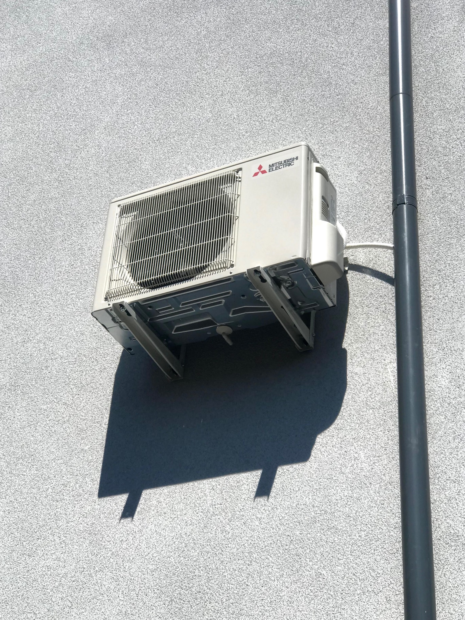 Zewnętrzna jednostka klimatyzacji Mitsubishi Electric zamontowana na szarej, chropowatej ścianie budynku, obok pionowej rury spustowej.