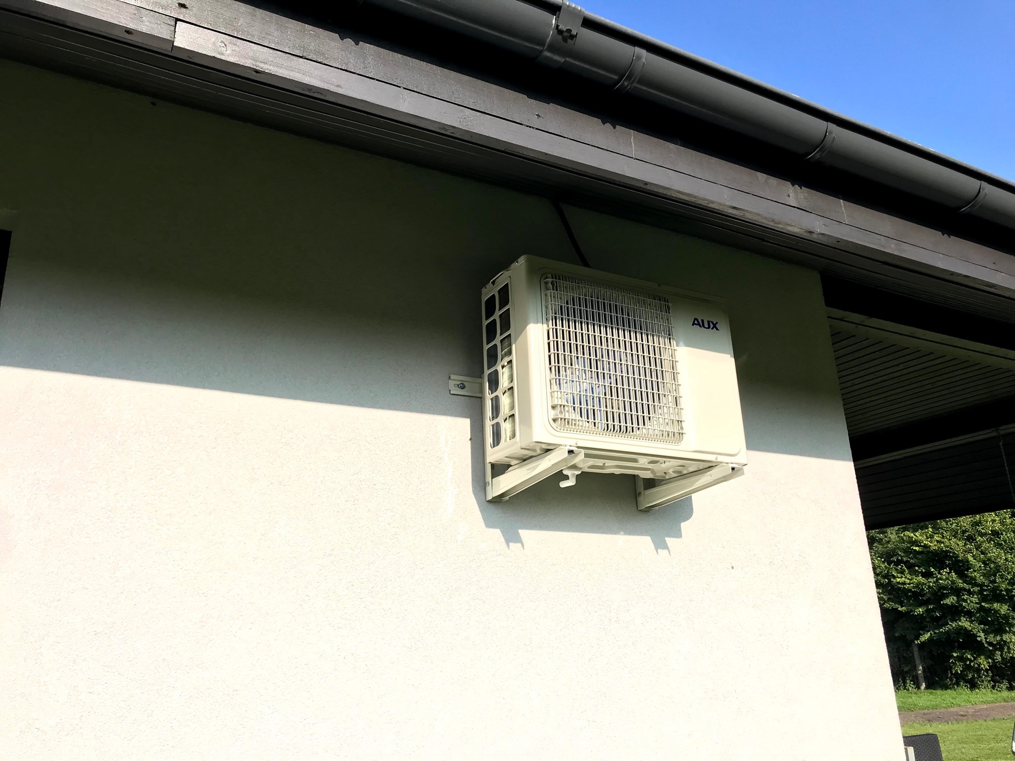 Zamontowana jednostka zewnętrzna klimatyzacji AUX na jasnej elewacji domu, widoczne rury spustowe i fragment ogrodu w tle.