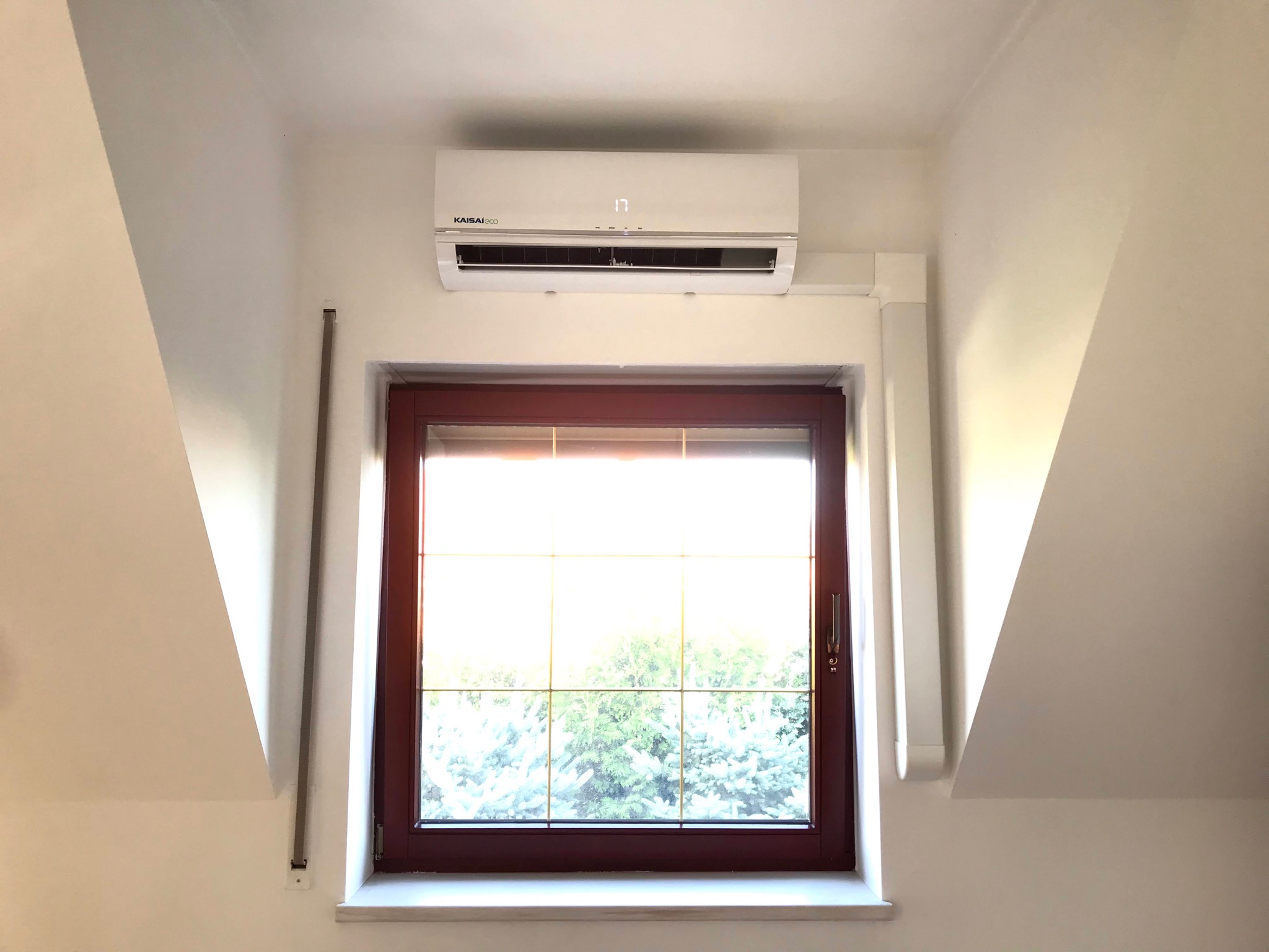 Zamontowany biały klimatyzator Kaisai Eco nad oknem z brązową ramą na poddaszu ze skośnymi ścianami, widok na zieleń za oknem.