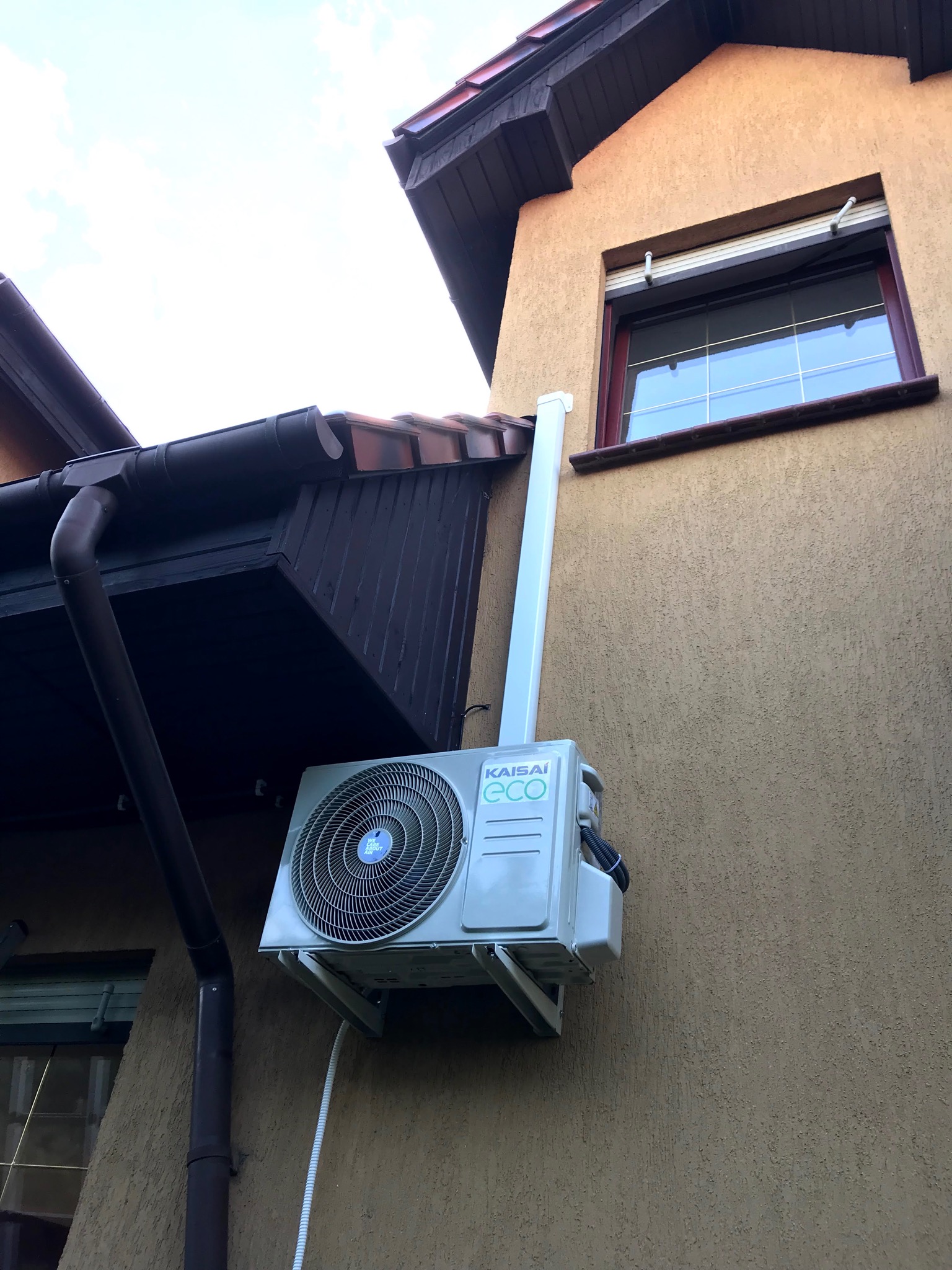 Zewnętrzna jednostka klimatyzacji Kaisai Eco zamontowana na elewacji domu z widocznymi rurami odprowadzającymi skropliny i przewodem zasilającym.