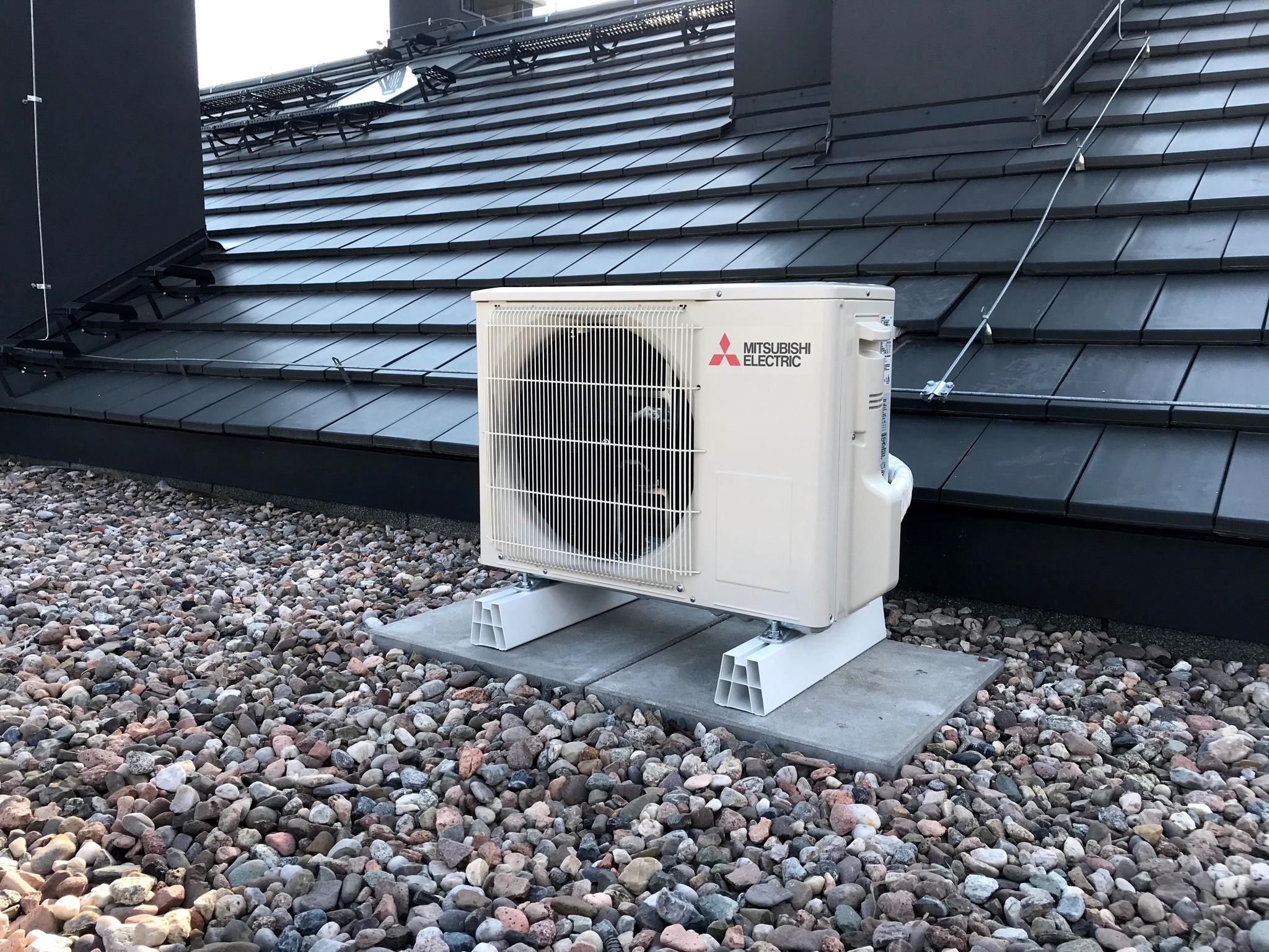 Zewnętrzna jednostka klimatyzacji Mitsubishi Electric zamontowana na betonowym fundamencie, otoczona kamienistym podłożem, na tle ciemnej, skośnej powierzchni dachu z widocznymi elementami...