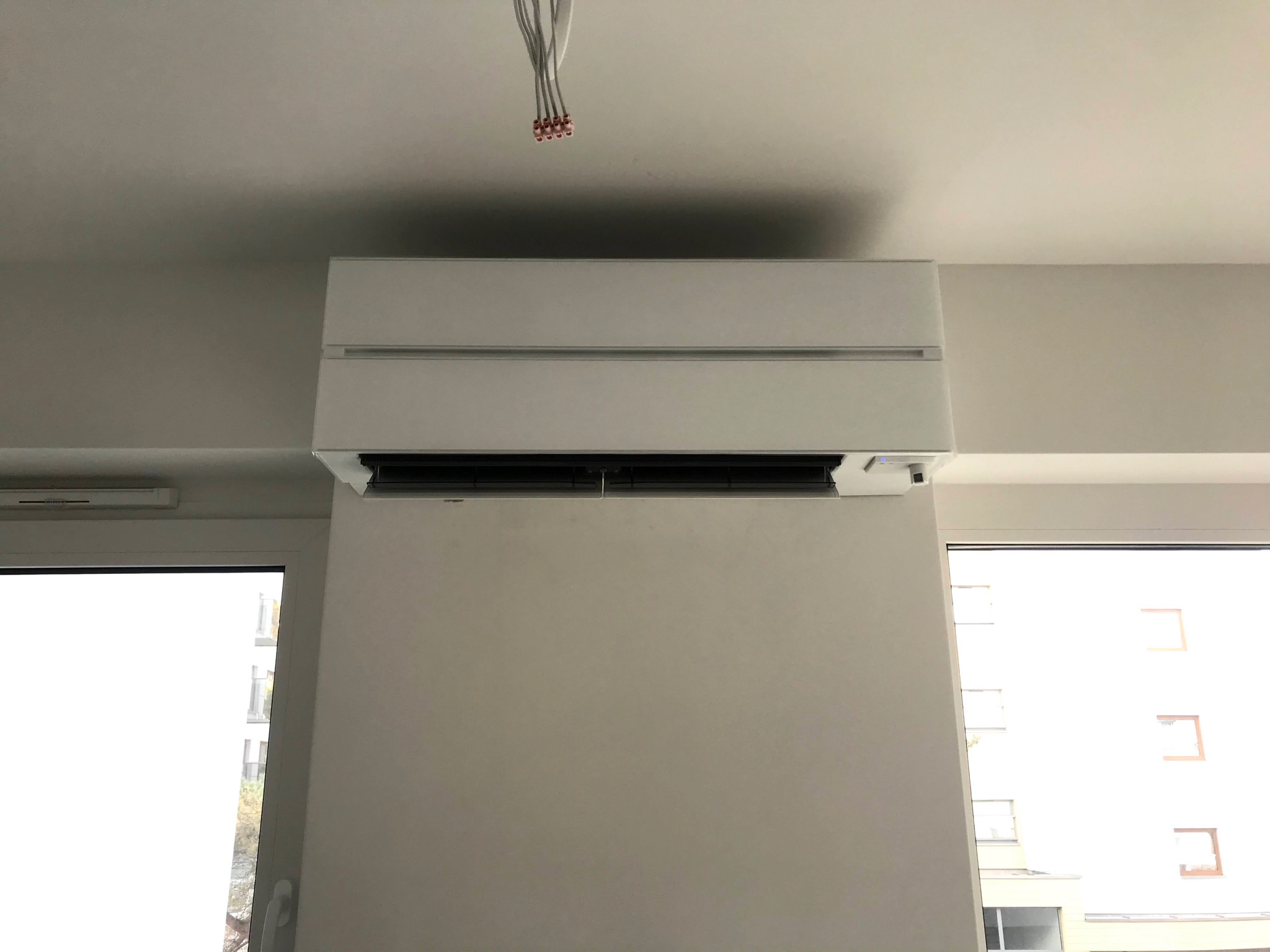 Nowoczesny, biały klimatyzator ścienny zamontowany w jasnym pomieszczeniu, z widocznymi przewodami elektrycznymi wystającymi z sufitu, sugerującymi świeżą instalację, z oknami po bokach kadru.