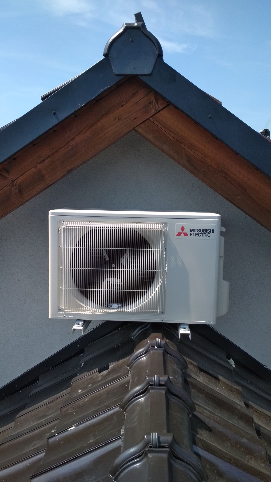 Zewnętrzna jednostka klimatyzatora Mitsubishi Electric zamontowana na wspornikach na dachu pokrytym ciemnobrązową dachówką ceramiczną, widok z dołu na tle szarej ściany i fragmentu drewnianej...