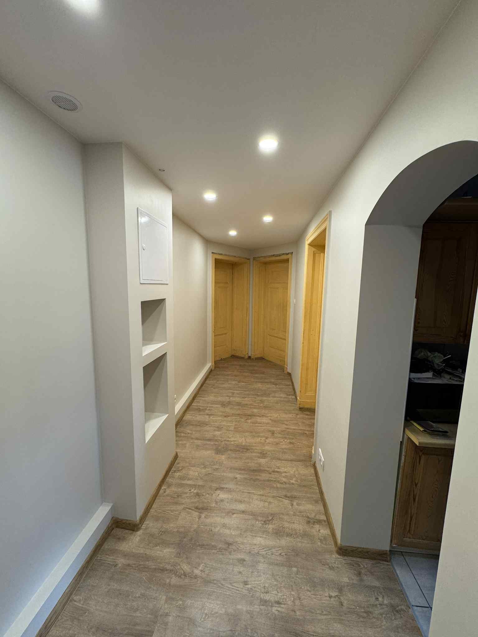 Korytarz po remoncie: świeżo pomalowane ściany w odcieniach szarości, drewnopodobna podłoga, wnęka w ścianie z półkami i widoczne drzwi.