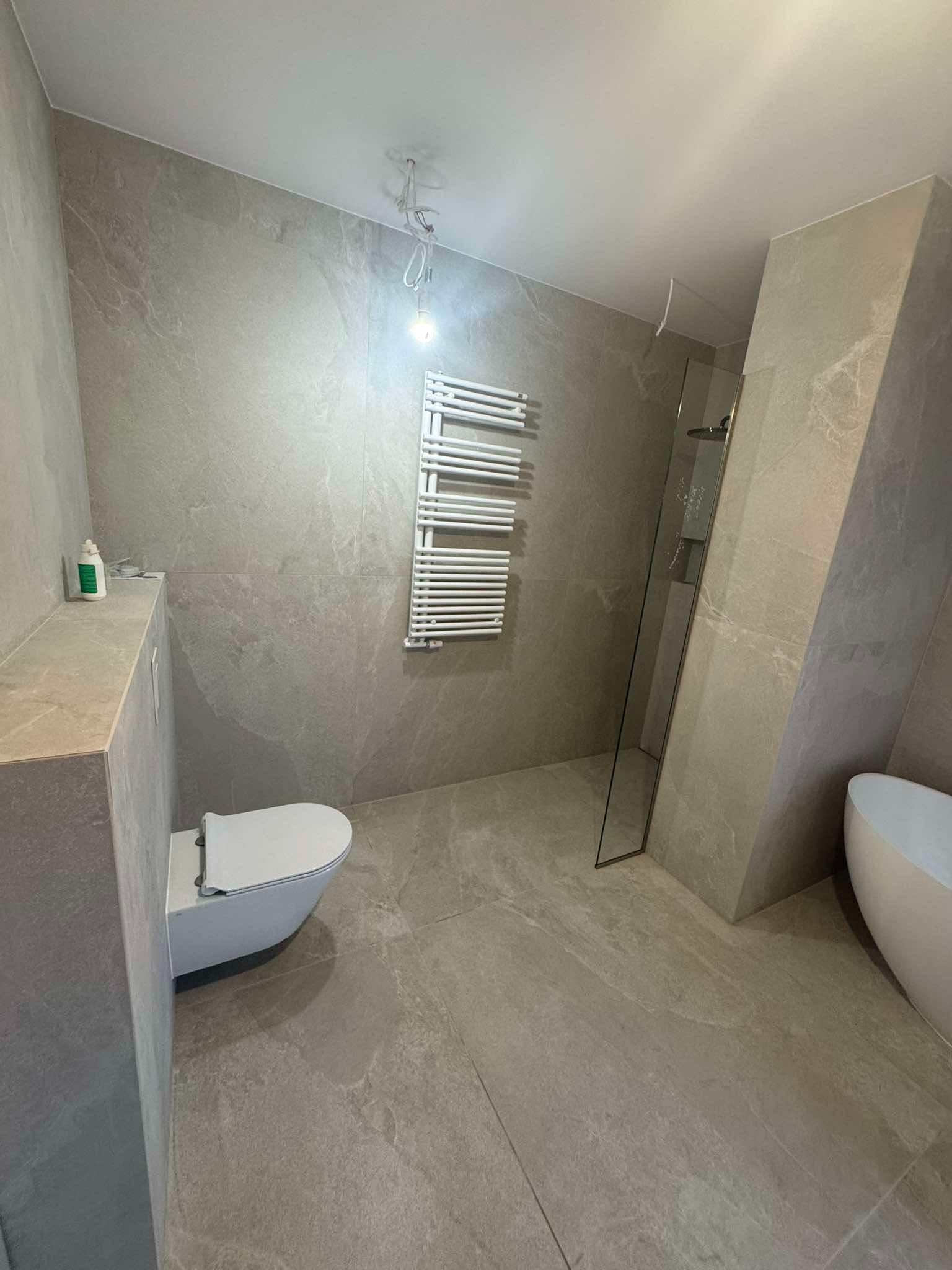 Wykończona łazienka w szarościach: płytki imitujące kamień na ścianach i podłodze, minimalistyczna toaleta, grzejnik drabinkowy i kabina prysznicowa ze szklaną ścianą.