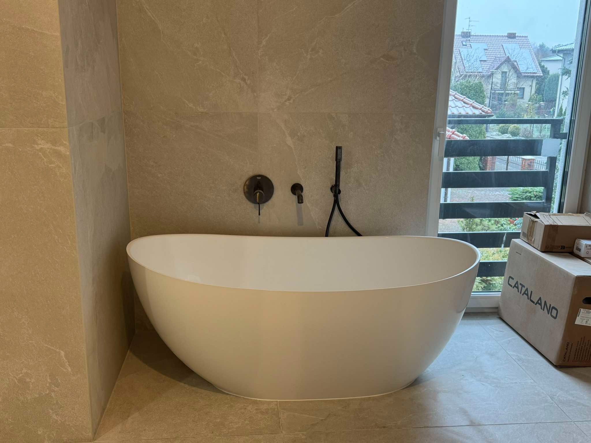 Nowoczesna, wolnostojąca wanna Catalano w łazience wykończonej płytkami imitującymi kamień, z widokiem na zewnątrz przez okno z żaluzjami.