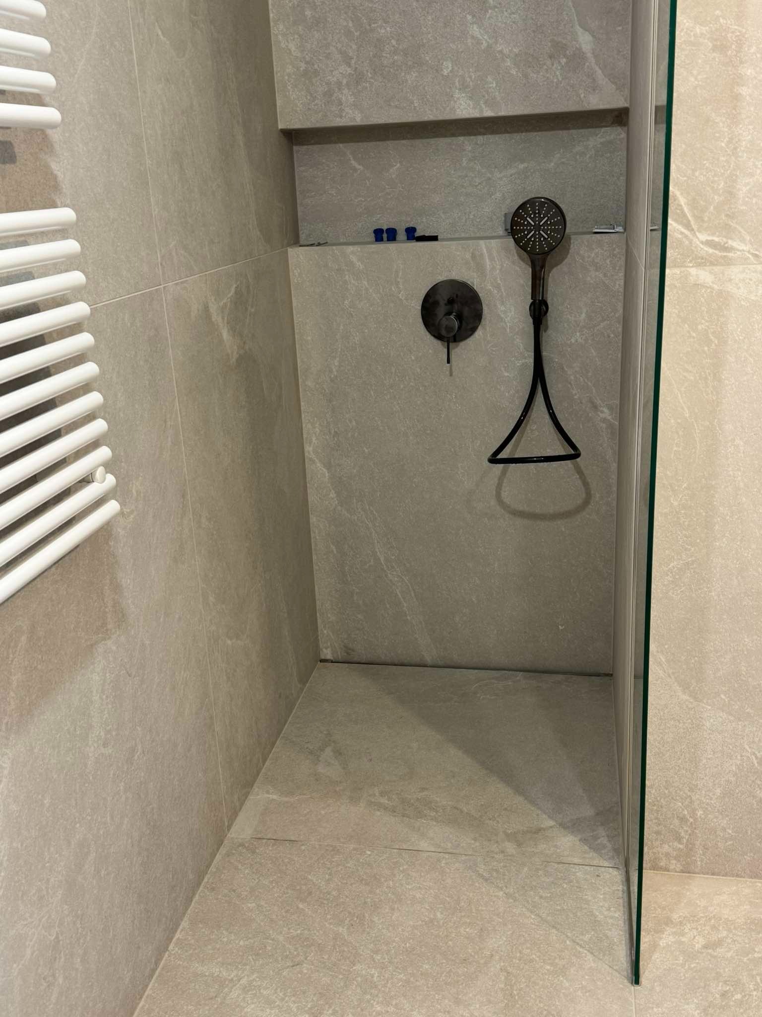 Wykończona kabina prysznicowa z szarymi płytkami imitującymi kamień, z czarną armaturą i grzejnikiem drabinkowym po lewej stronie. Minimalistyczny design.