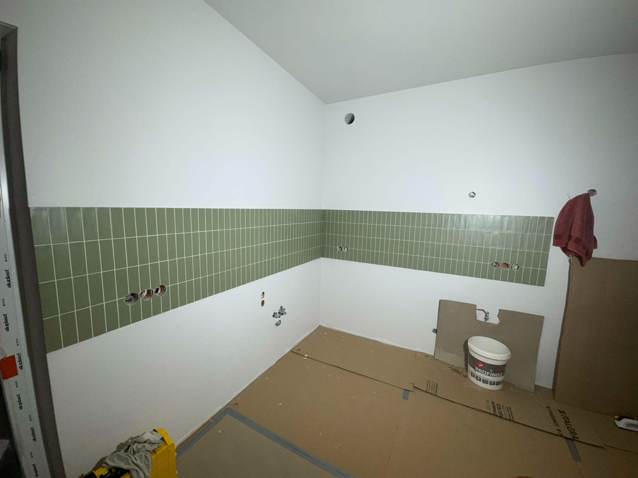 Fragment łazienki w trakcie prac wykończeniowych: zielone płytki ułożone do połowy ściany, białe ściany powyżej, widoczne instalacje, wiadro z zaprawą.