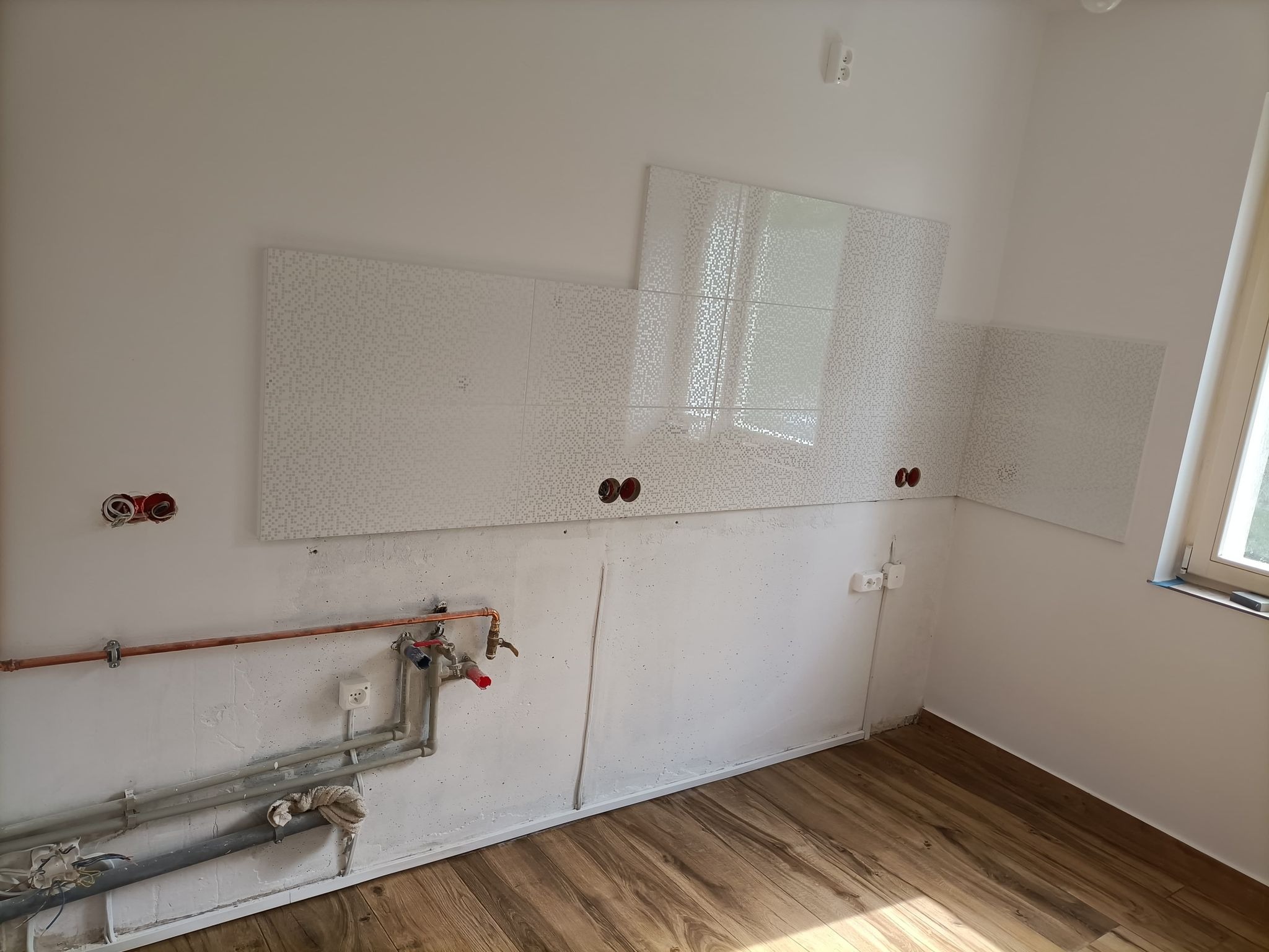 Fragment ściany w trakcie prac wykończeniowych w łazience: częściowo ułożone białe płytki z delikatnym wzorem, odsłonięte instalacje wodne i elektryczne, drewnopodobna podłoga, okno z widocznym...