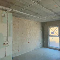 Surowy stan wnętrza z widoczną instalacją elektryczną: kable w białych listwach prowadzone po suficie i ścianach z betonu komórkowego, przygotowane punkty pod gniazdka i włączniki, okno...