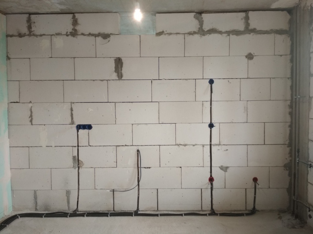 Surowa ściana z betonu komórkowego z widocznymi przewodami elektrycznymi ułożonymi wzdłuż podłogi i pionowo w górę, zakończonymi puszkami instalacyjnymi w kolorze niebieskim i czerwonym.