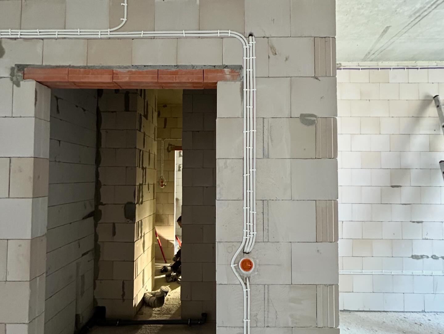 Białe rurki instalacji elektrycznej prowadzone po ścianie z bloczków betonu komórkowego, widoczny otwór drzwiowy z nadprożem z cegły, w tle drabina i dalsza część instalacji.